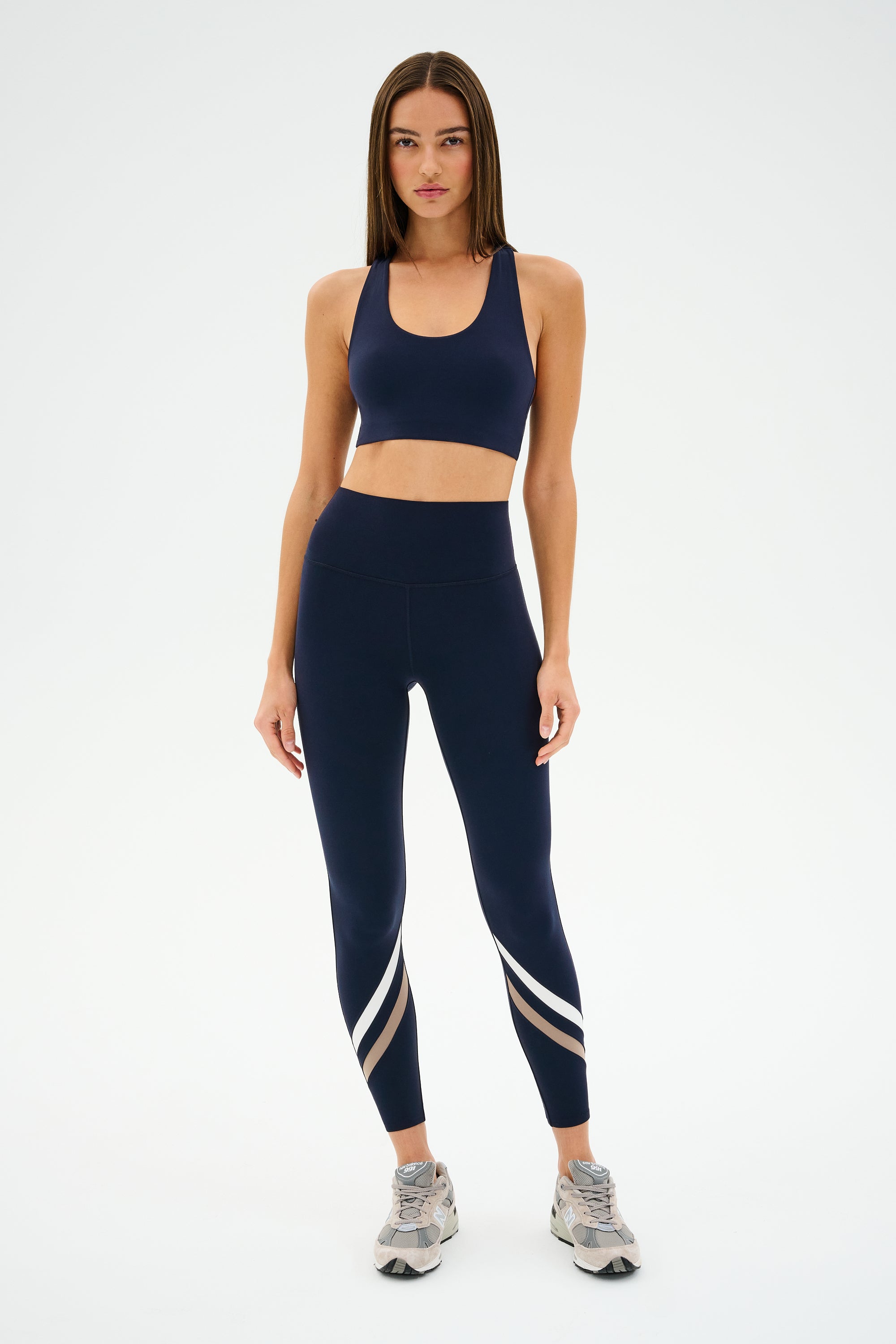 A person in navy athletic wear stands against a plain background, donning SPLITS59's Chevron Airweight High Waist 7/8 leggings in Indigo/Latte, paired with a sports bra and sleek sneakers.