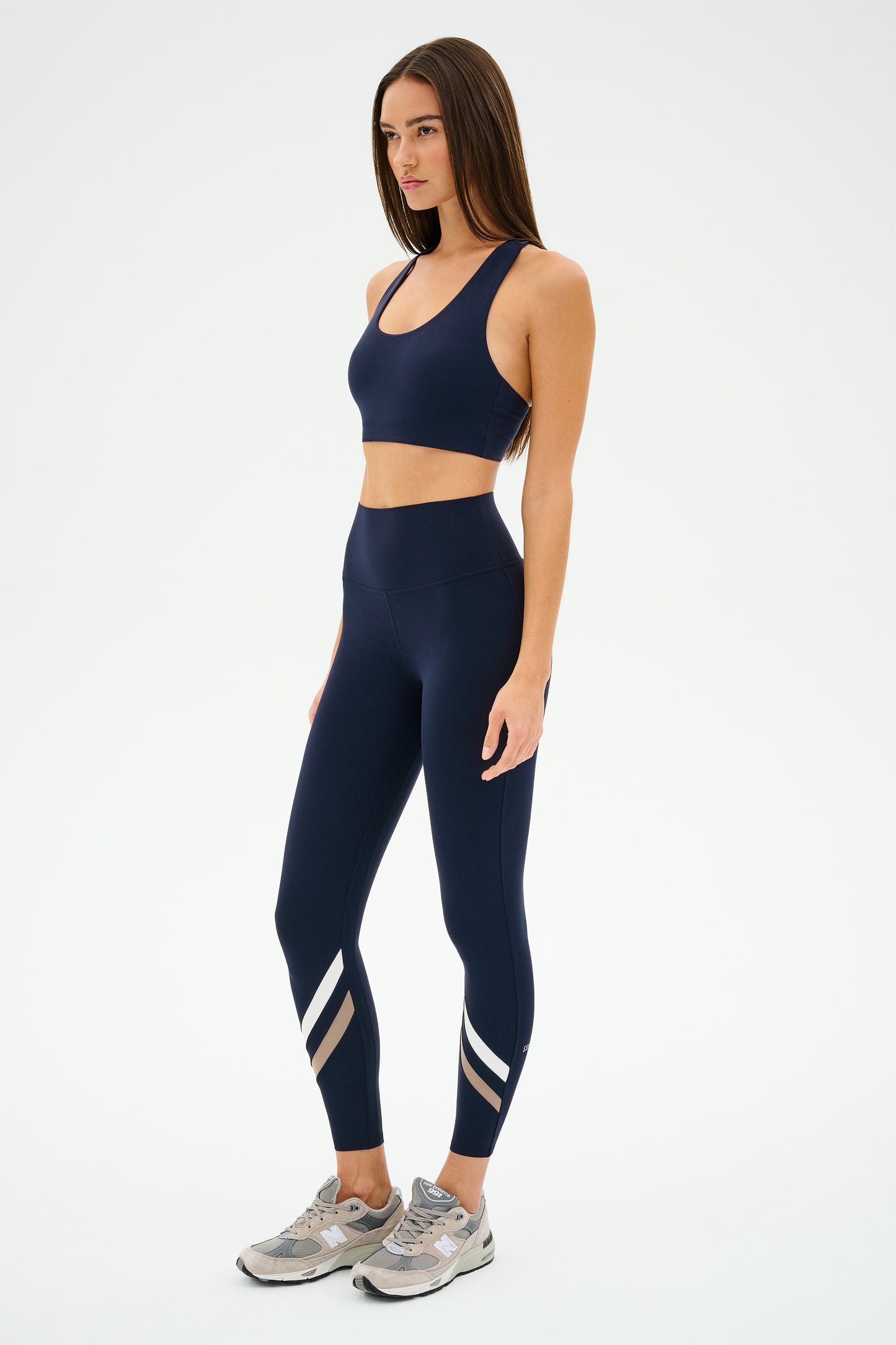 Against a plain background, someone wears SPLITS59's Chevron Airweight High Waist 7/8 leggings in Indigo/Latte with a dark blue sports bra and sneakers.