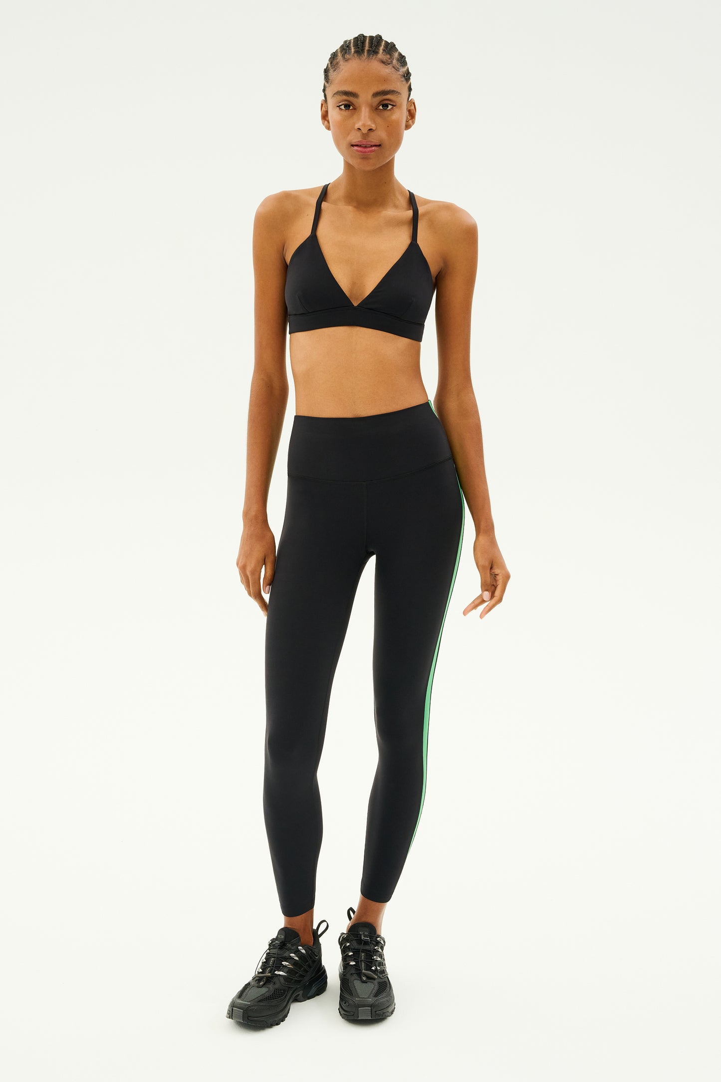 A person wearing the SPLITS59 Theo Rigor Bra in black, part of an ultra-luxe athletic outfit, paired with green-striped leggings and black sneakers, stands on a simple white background.