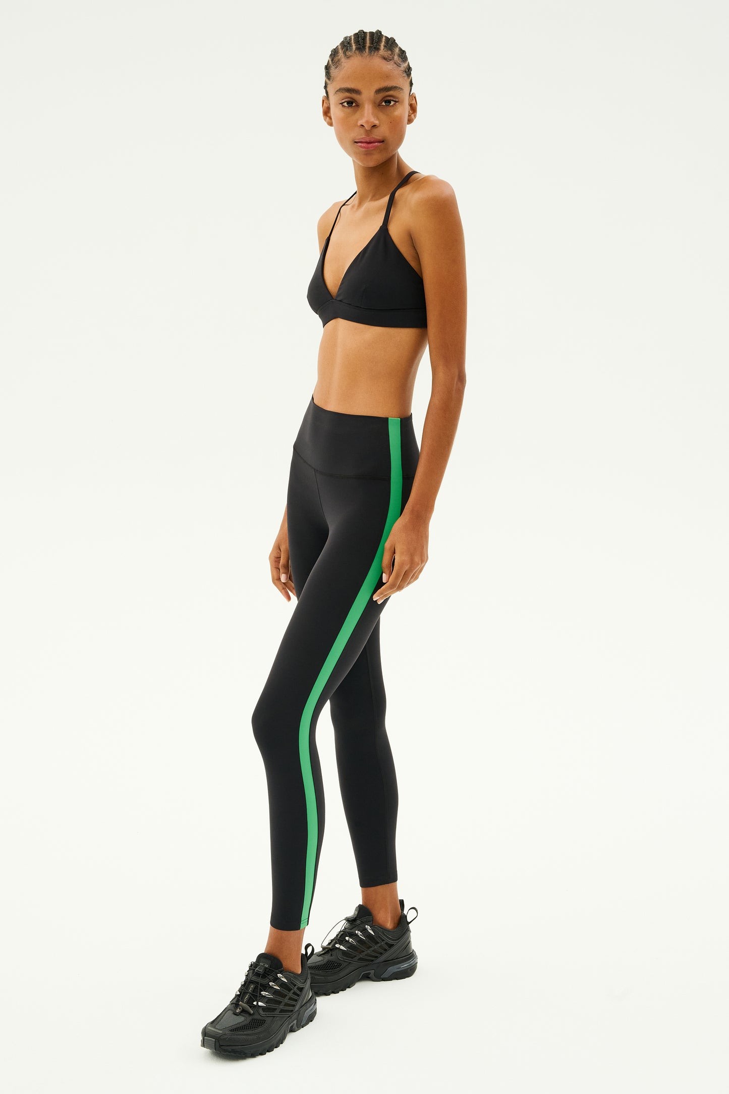 A person stands against a plain background, wearing the stylish Theo Rigor Bra - Black by SPLITS59, paired with black leggings that feature green stripes. The outfit highlights multi-sport performance and is completed with sleek black sneakers, all made from ultra-luxe Rigor fabric for exceptional comfort and style.