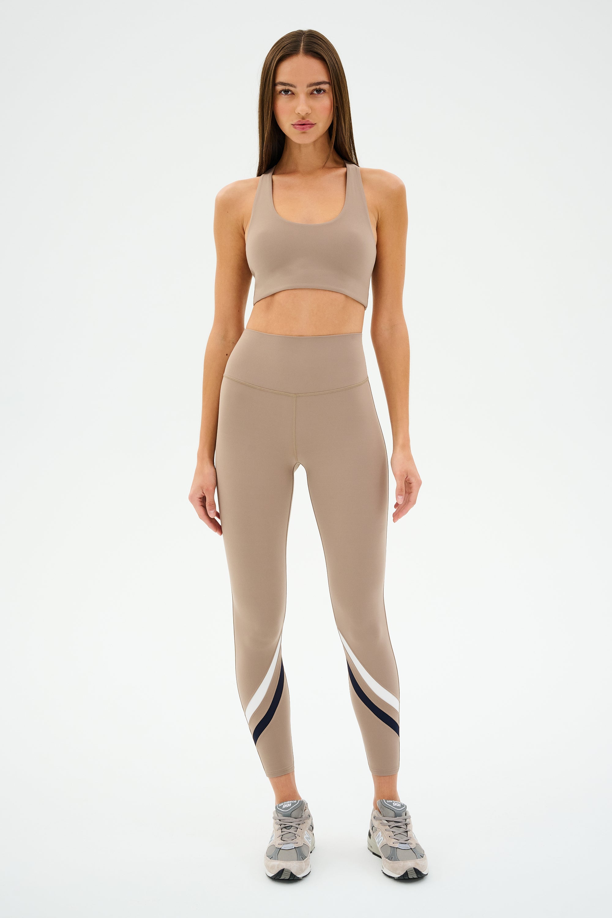 A woman poses in a SPLITS59 Sara Airweight Bra - Latte and beige leggings featuring stripe details, paired with running shoes, against a plain background. This ultra-luxe outfit is perfect for hot yoga, seamlessly blending style and function.