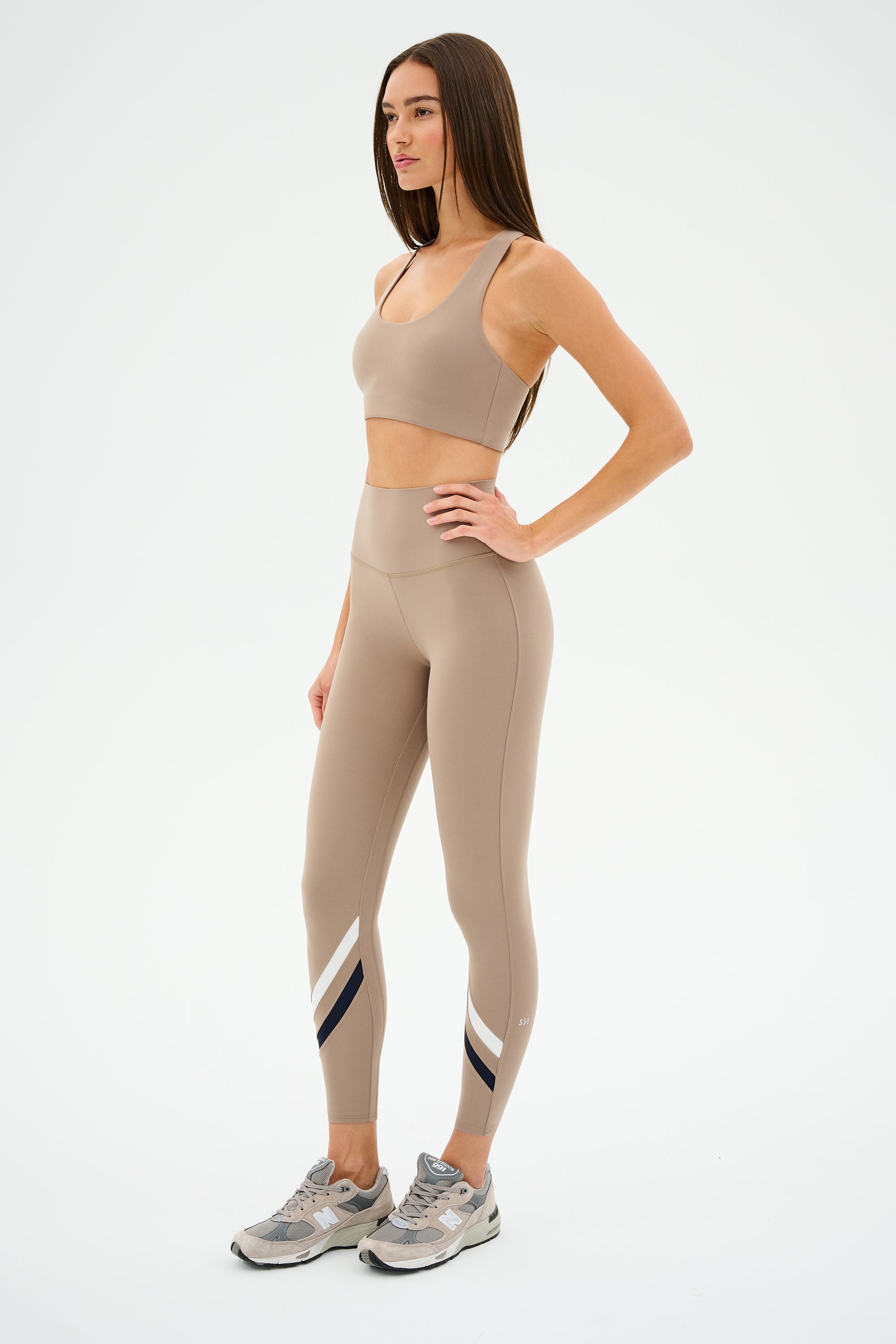 Dressed in ultra-luxe athletic wear, a person stands in profile wearing a SPLITS59 Sara Airweight Bra in Latte and matching sneakers against a plain background.