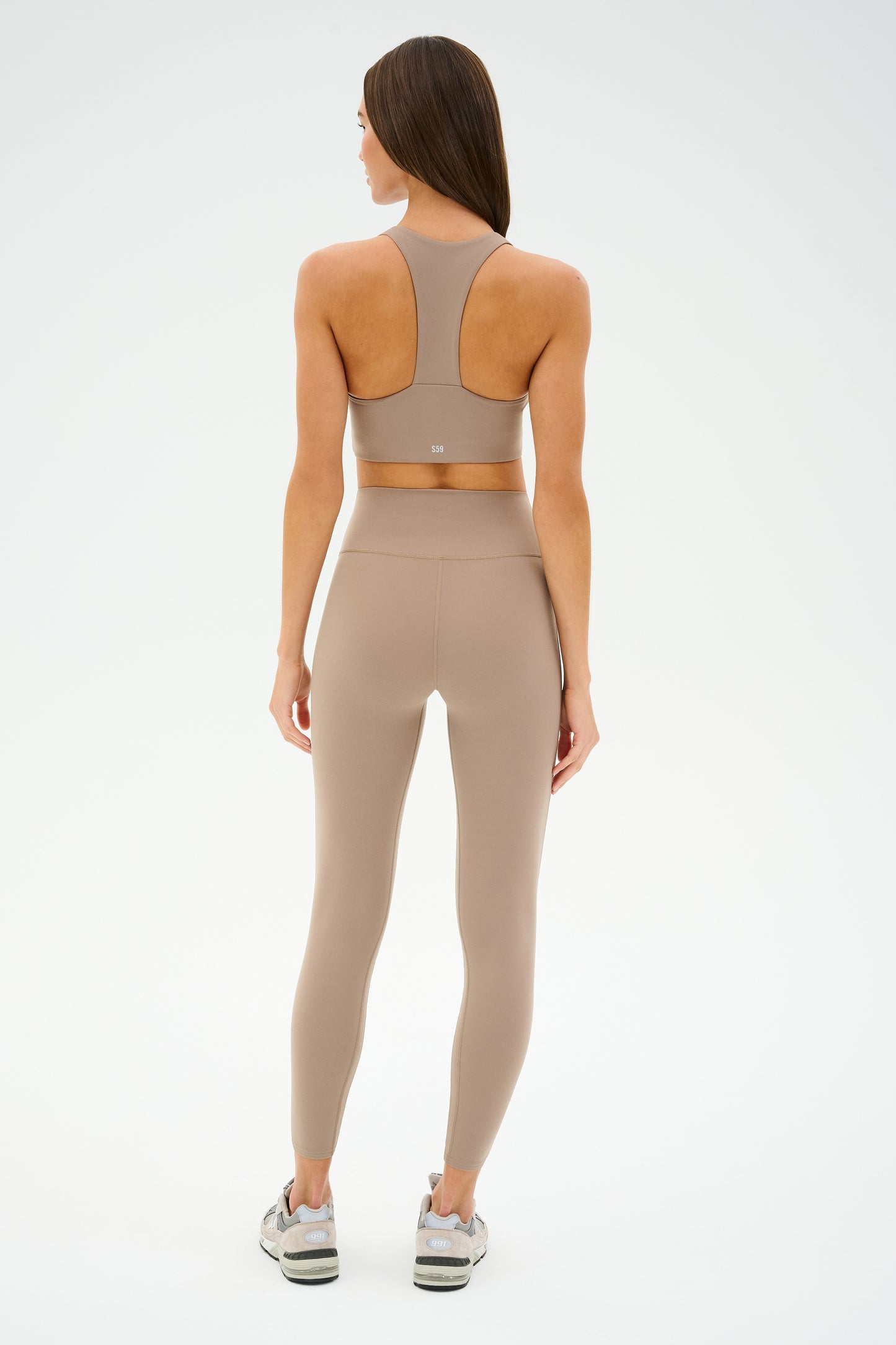 A woman in ultra-luxe beige sportswear and sneakers, featuring the SPLITS59 Sara Airweight Bra in Latte, stands with her back to the camera against a plain background.