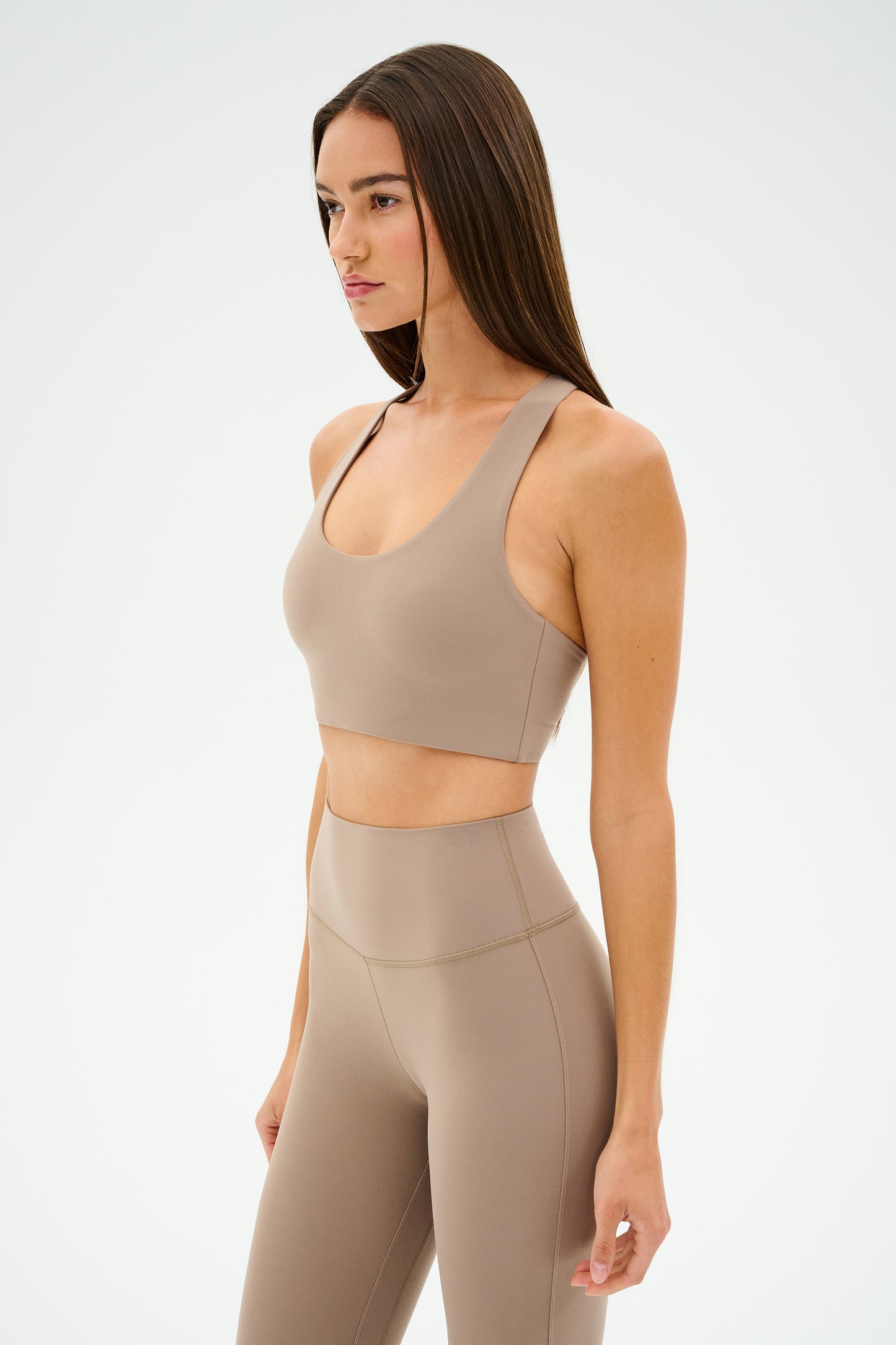 A person with long hair, wearing a Latte-colored SPLITS59 Sara Airweight Bra and matching leggings, stands against a plain background exuding an ultra-luxe vibe, ready for hot yoga.