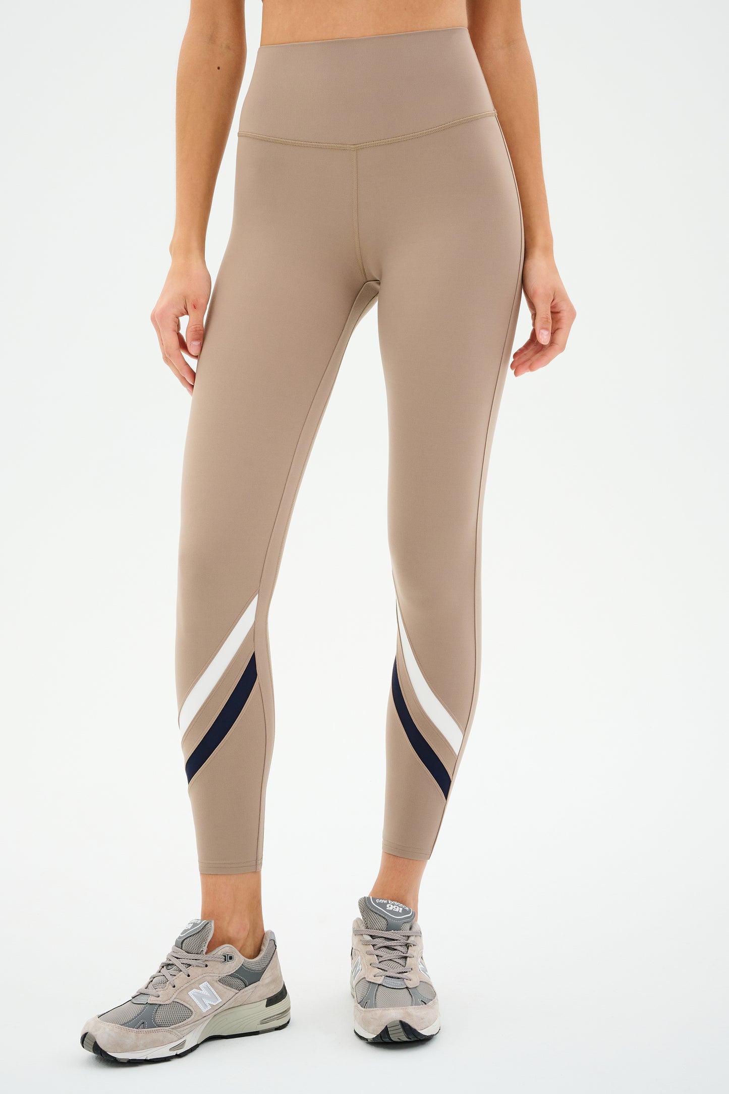 Against a plain background, an individual is wearing SPLITS59's Chevron Airweight High Waist 7/8 leggings in Latte/Indigo, featuring ultra luxe fabric with white and black stripes, paired seamlessly with gray sneakers.