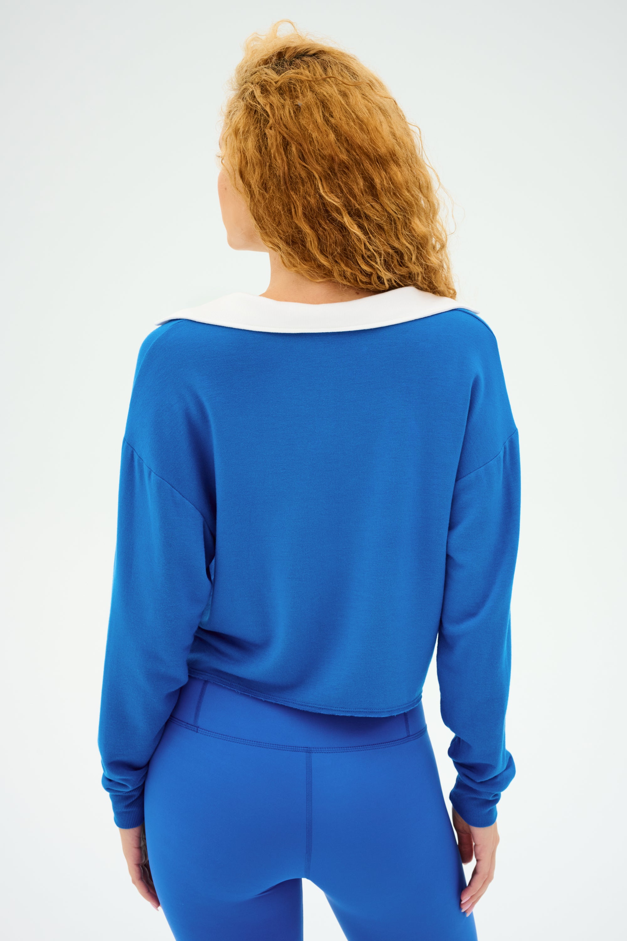 A person with curly hair, clad in a SPLITS59 Joan Fleece Polo Sweatshirt in Classic Blue/White and leggings, stands facing away against a plain background.