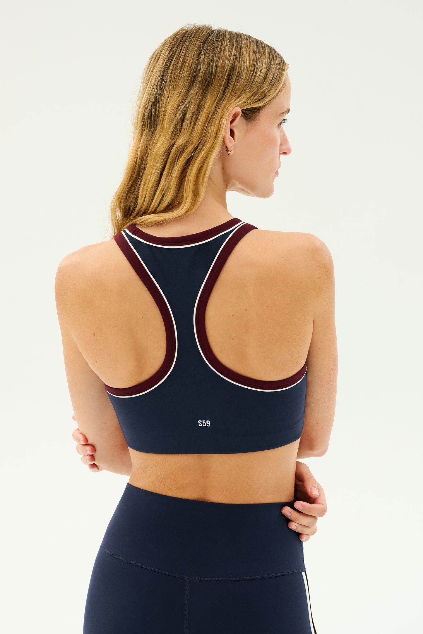 A woman with blonde hair is standing with her back to the camera, wearing an indigo and dark cherry racerback bra, known as the Robbie Airweight Bra by SPLITS59, paired with matching ultra luxe Airweight high-waisted leggings.