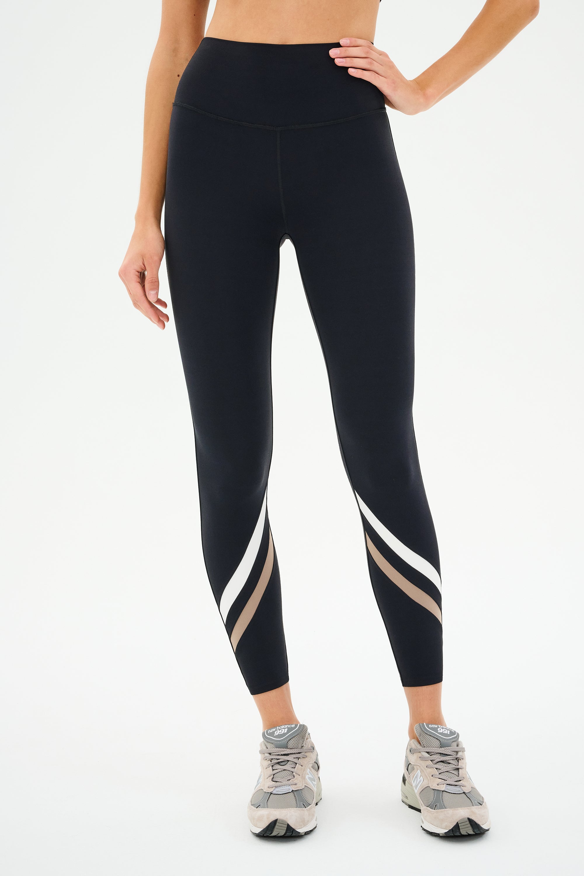 Someone is shown from the waist down wearing SPLITS59 Chevron Airweight High Waist 7/8 leggings in Black/Latte, featuring white and brown stripes near the ankles, paired with gray sneakers against a plain background.