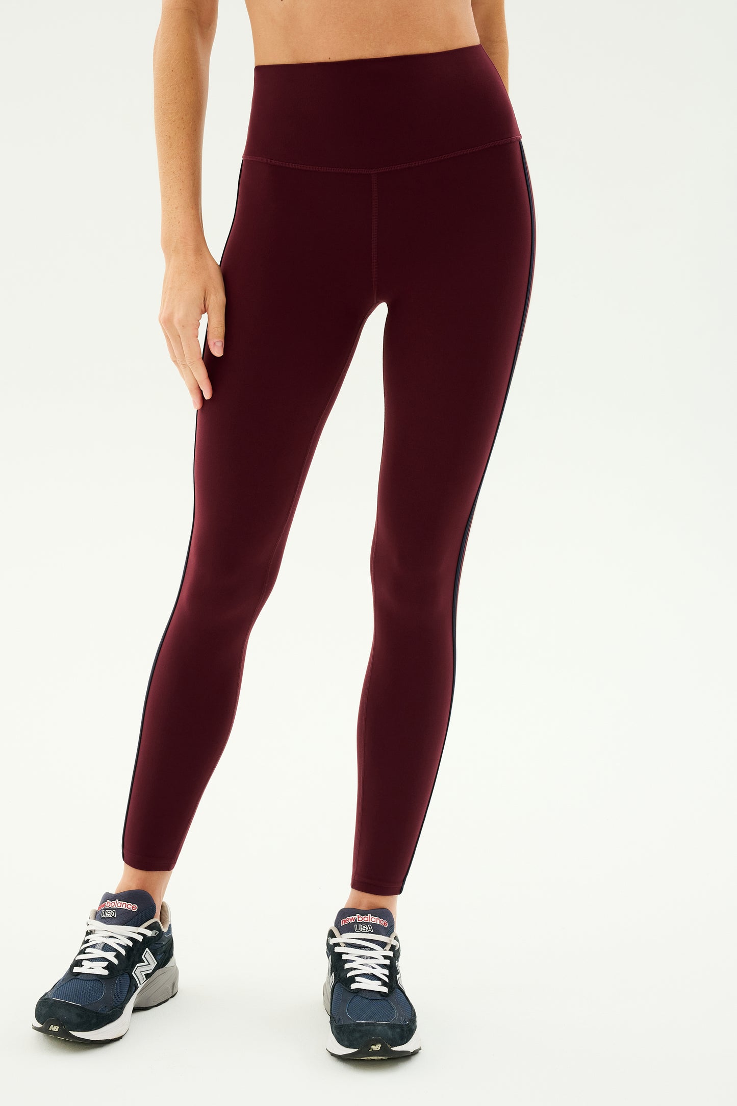 A person wearing SPLITS59's Ella High Waist Airweight 7/8 leggings in Dark Cherry/Indigo and navy sneakers stands against a plain background.