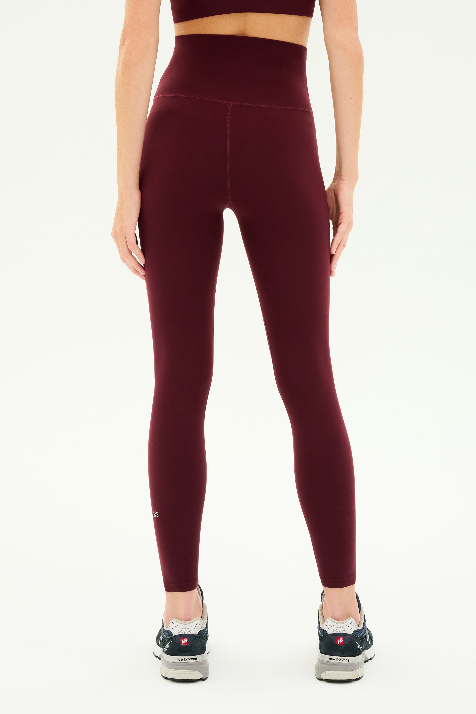 A person wearing SPLITS59's Airweight Super High Waist 7/8 leggings in Dark Cherry and grey sneakers is standing with their back facing the camera against a plain white background.