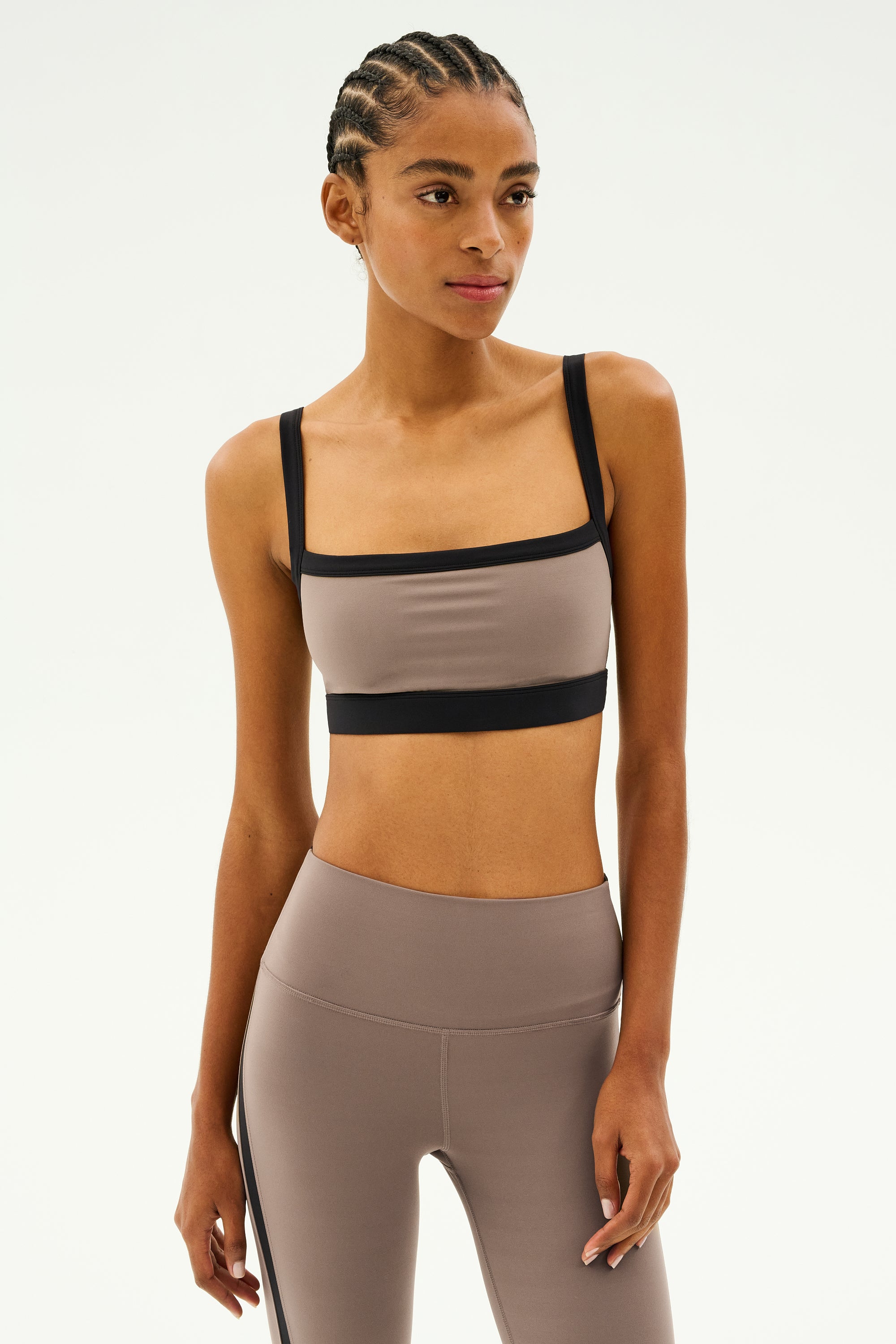 A person wearing a beige and black athletic outfit, featuring the stylish SPLITS59 Monah Rigor Bra in Lentil/Black, stands against a plain background.