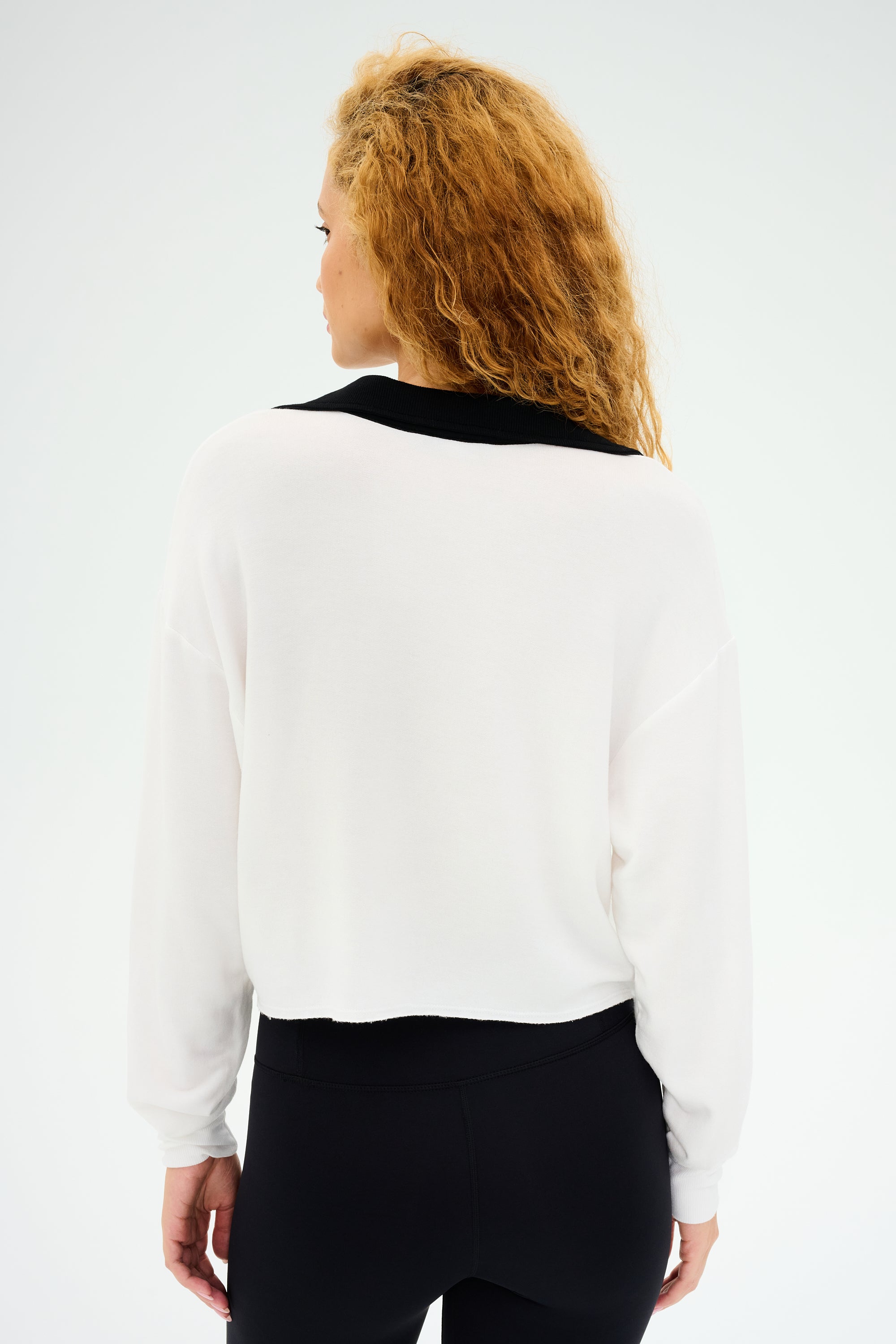 A person with curly hair, wearing the SPLITS59 Joan Fleece Polo Sweatshirt in White/Black and paired with black leggings, faces away from the camera.