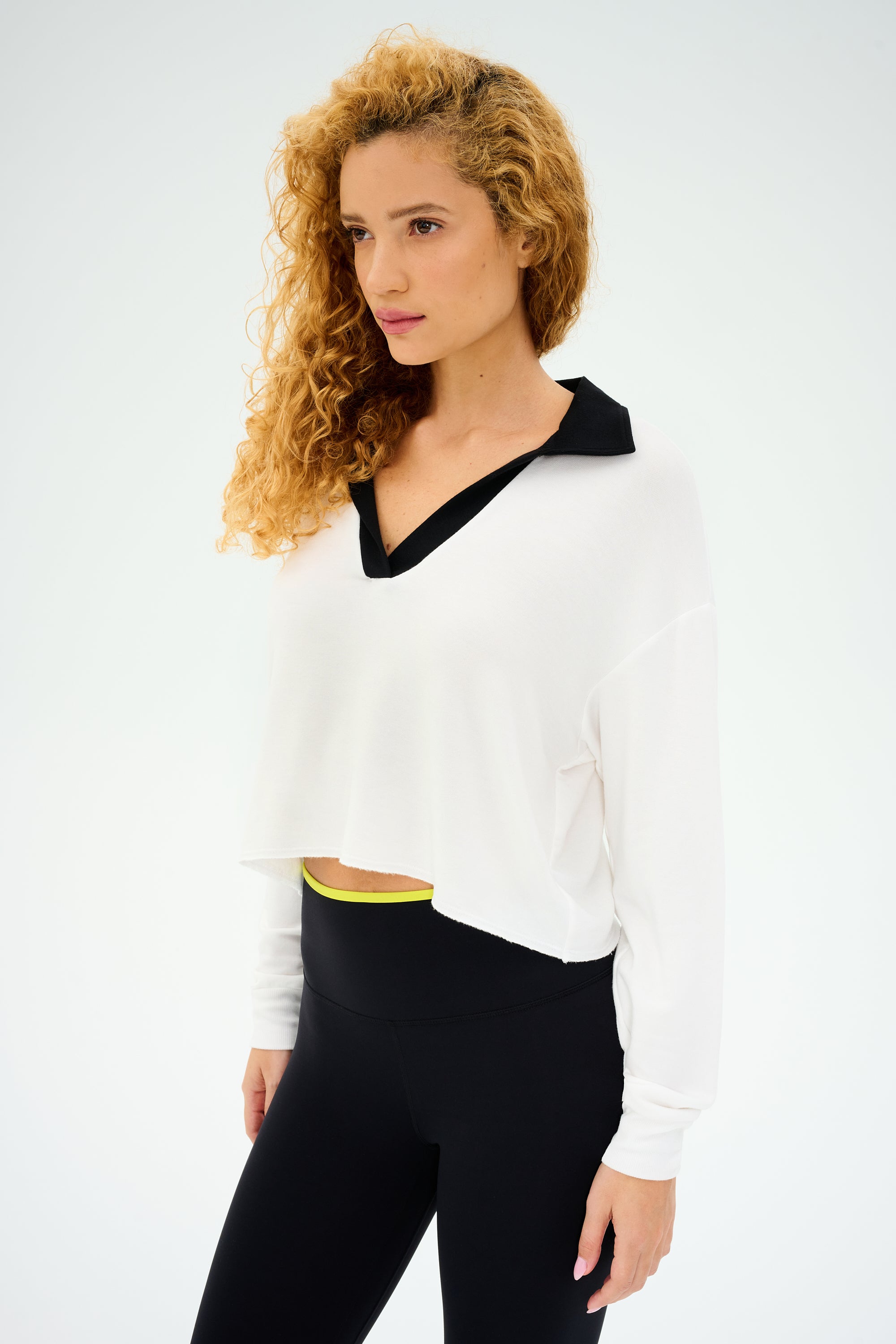 A curly-haired person is wearing a SPLITS59 Joan Fleece Polo Sweatshirt in white with a black collar and black leggings, standing against a plain background.