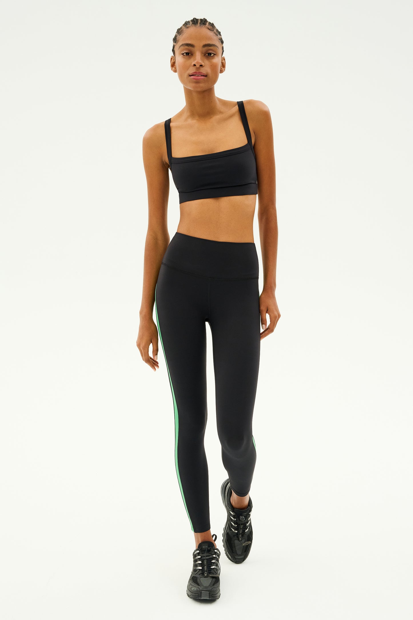 Wearing the Monah Rigor Bra in black by SPLITS59, a person strides confidently, enjoying solid support. They pair it with matching leggings that have a green stripe and complete the look with black sneakers, making it ideal for a workout set against a plain background.