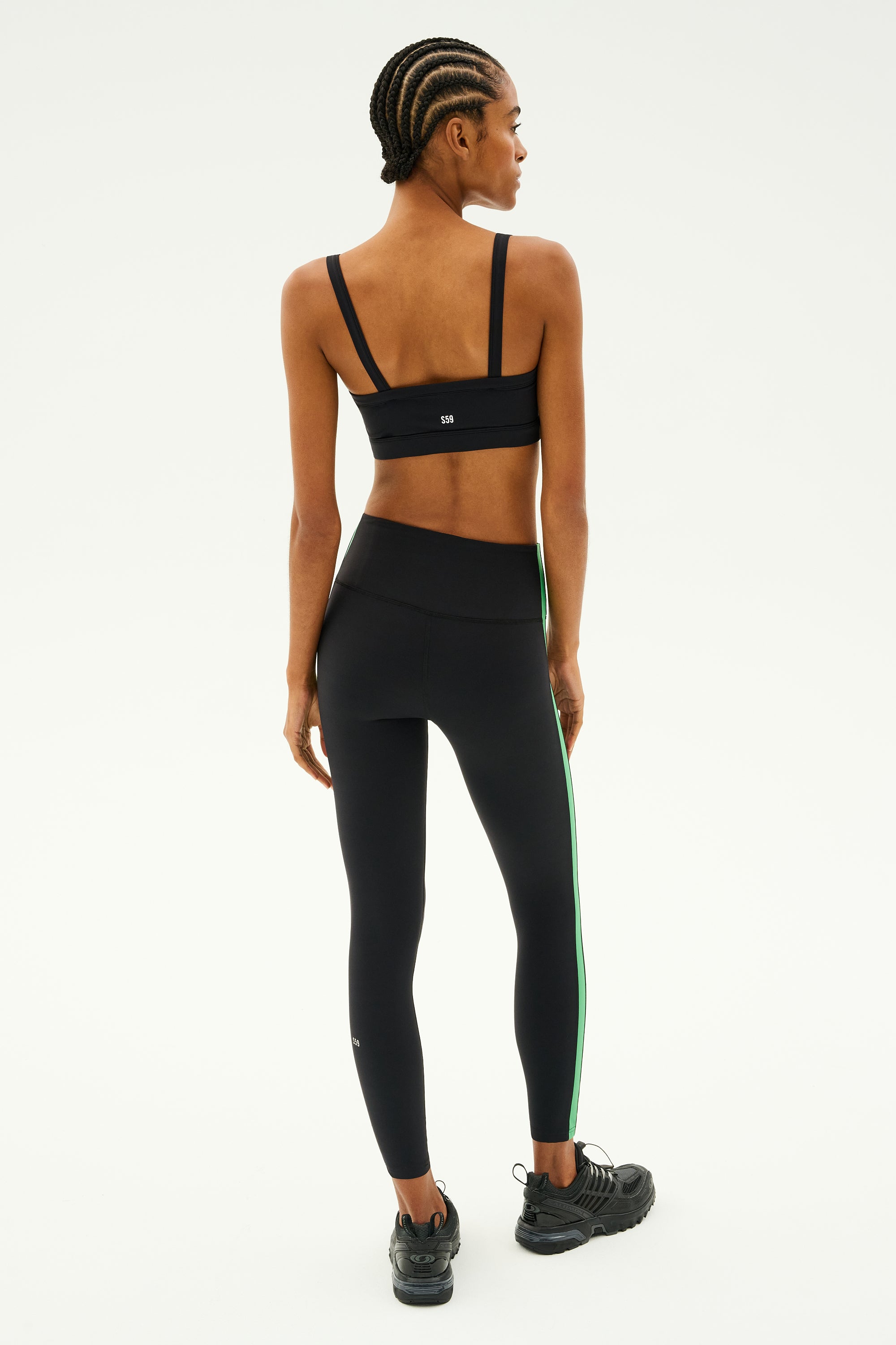 A person with braided hair stands facing away, ready for a workout. They're wearing the SPLITS59 Monah Rigor Bra in black for solid support, along with black leggings featuring a green stripe and black sneakers.