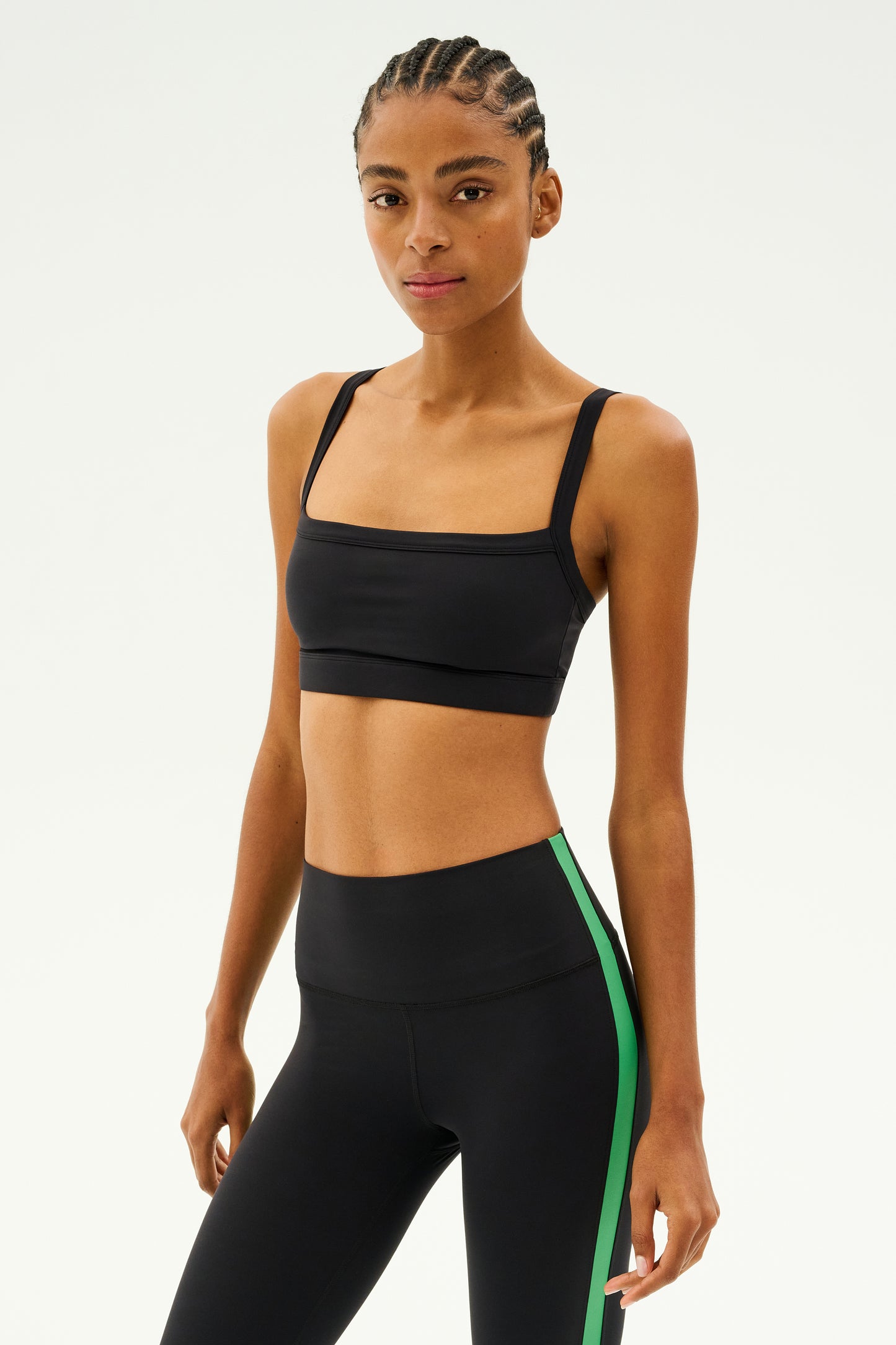 Adorned in the stylish Monah Rigor Bra - Black by SPLITS59, this person wears a black sports outfit accented with a fashionable green stripe, capturing a retro sport vibe. They pose assuredly against a simple backdrop, prepared to take on any workout challenge.