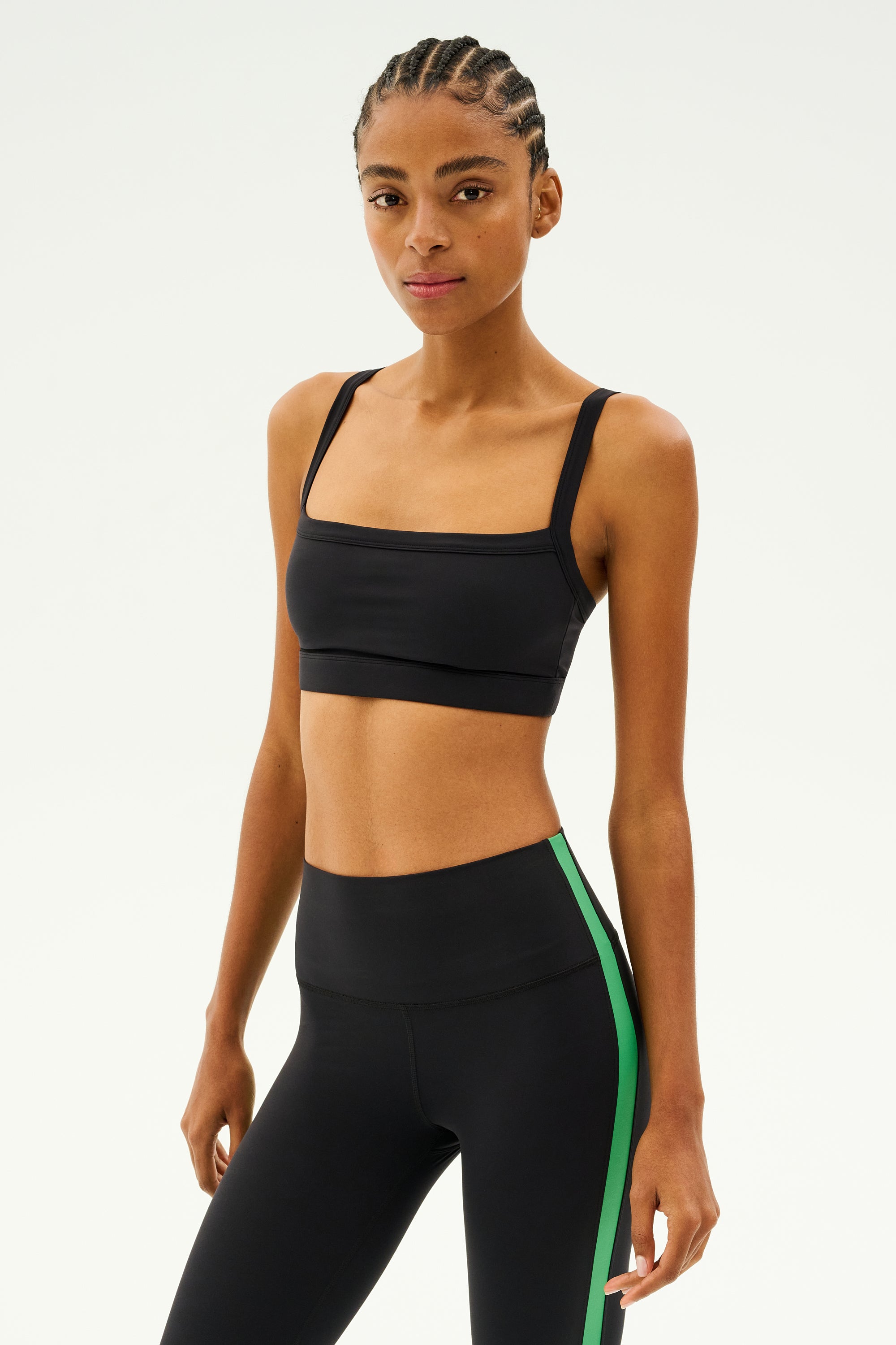 Adorned in the stylish Monah Rigor Bra - Black by SPLITS59, this person wears a black sports outfit accented with a fashionable green stripe, capturing a retro sport vibe. They pose assuredly against a simple backdrop, prepared to take on any workout challenge.