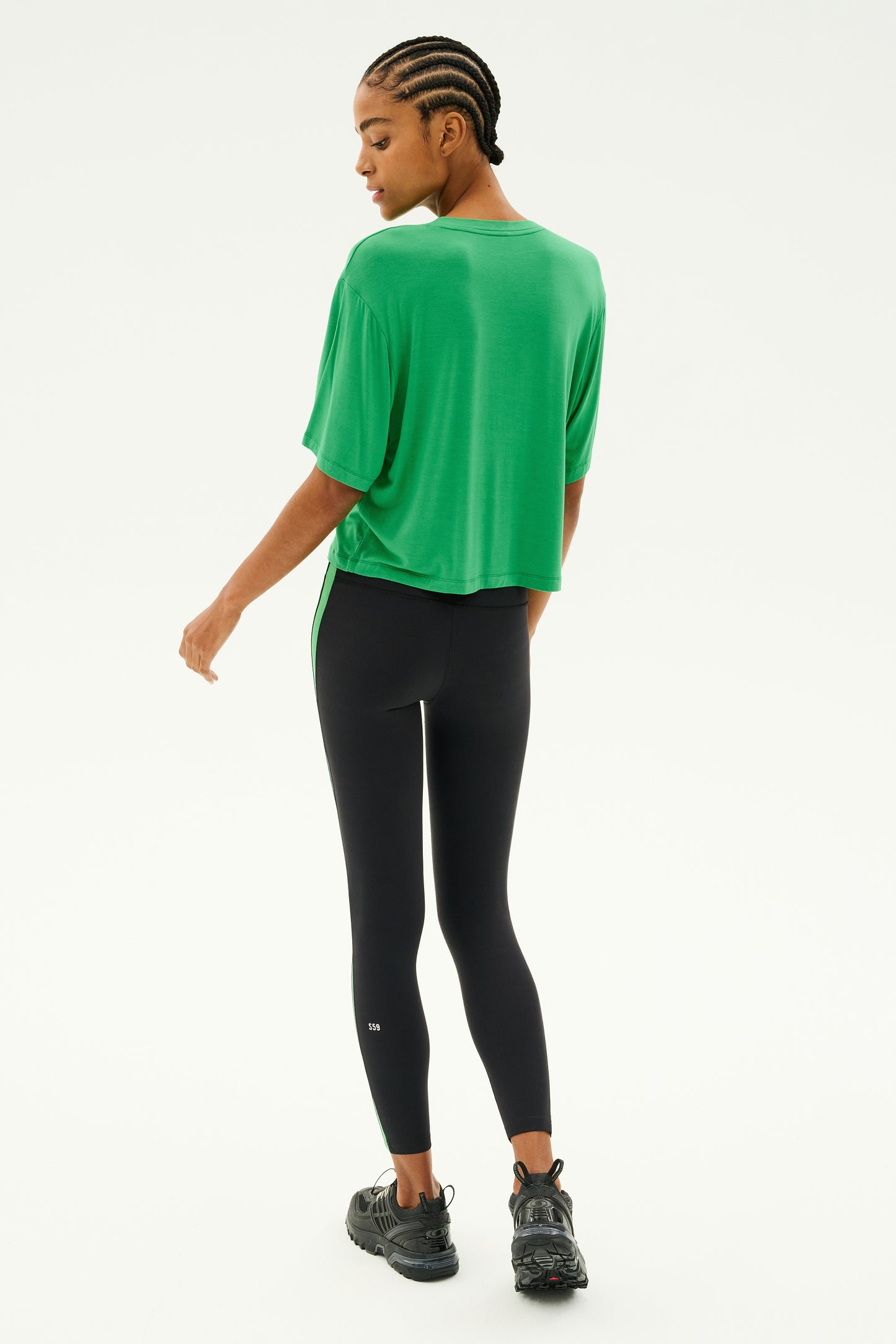 An individual in a SPLITS59 Djuna Oversized Jersey Tee in Grass and black leggings, facing sideways with black sneakers, gives the impression of being prepared for gym workouts, set against a plain white background.