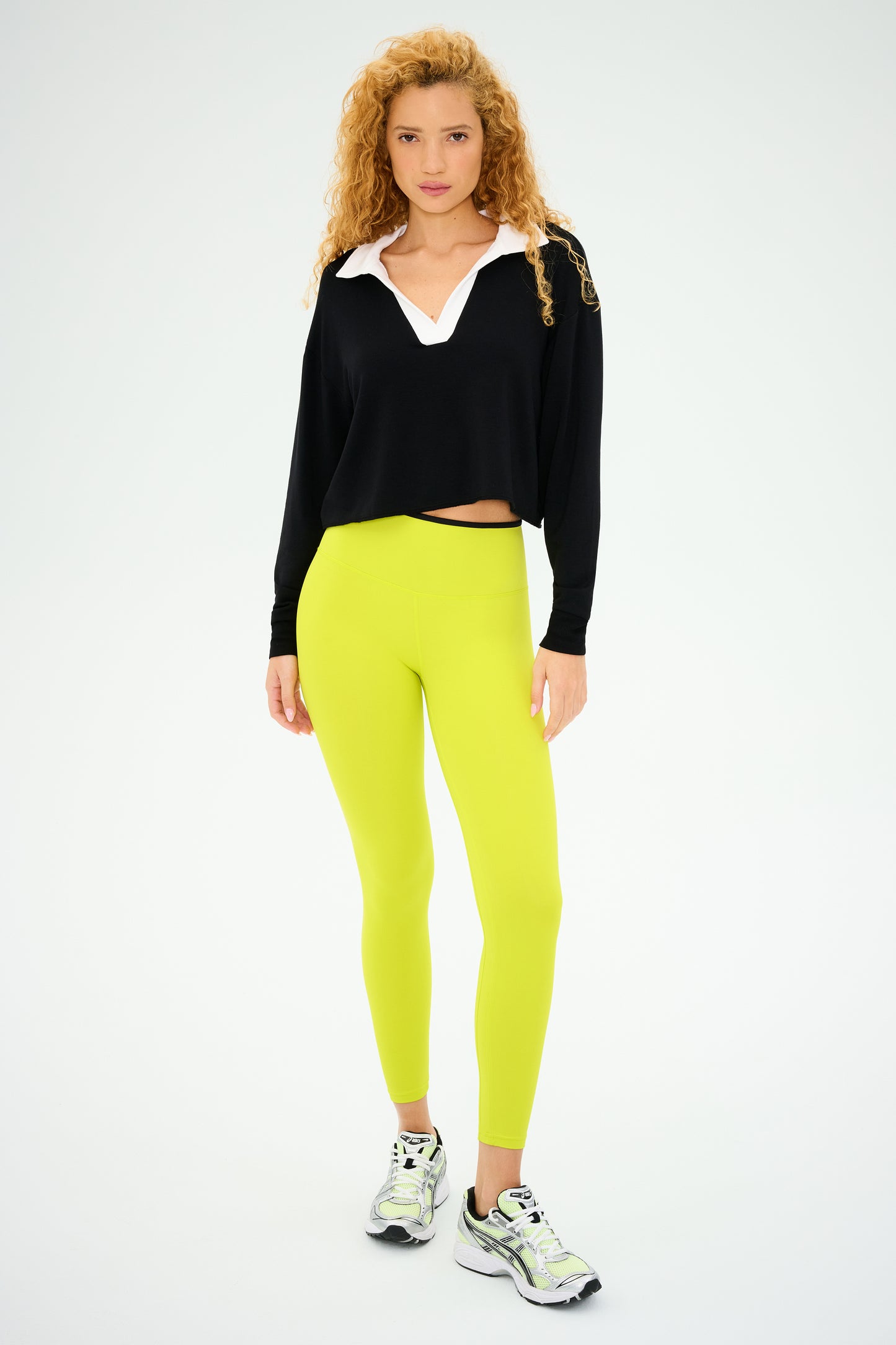 A person with curly hair stands confidently in SPLITS59's Joan Fleece Polo Sweatshirt in black/white and lime green leggings, paired with sneakers against a plain background.