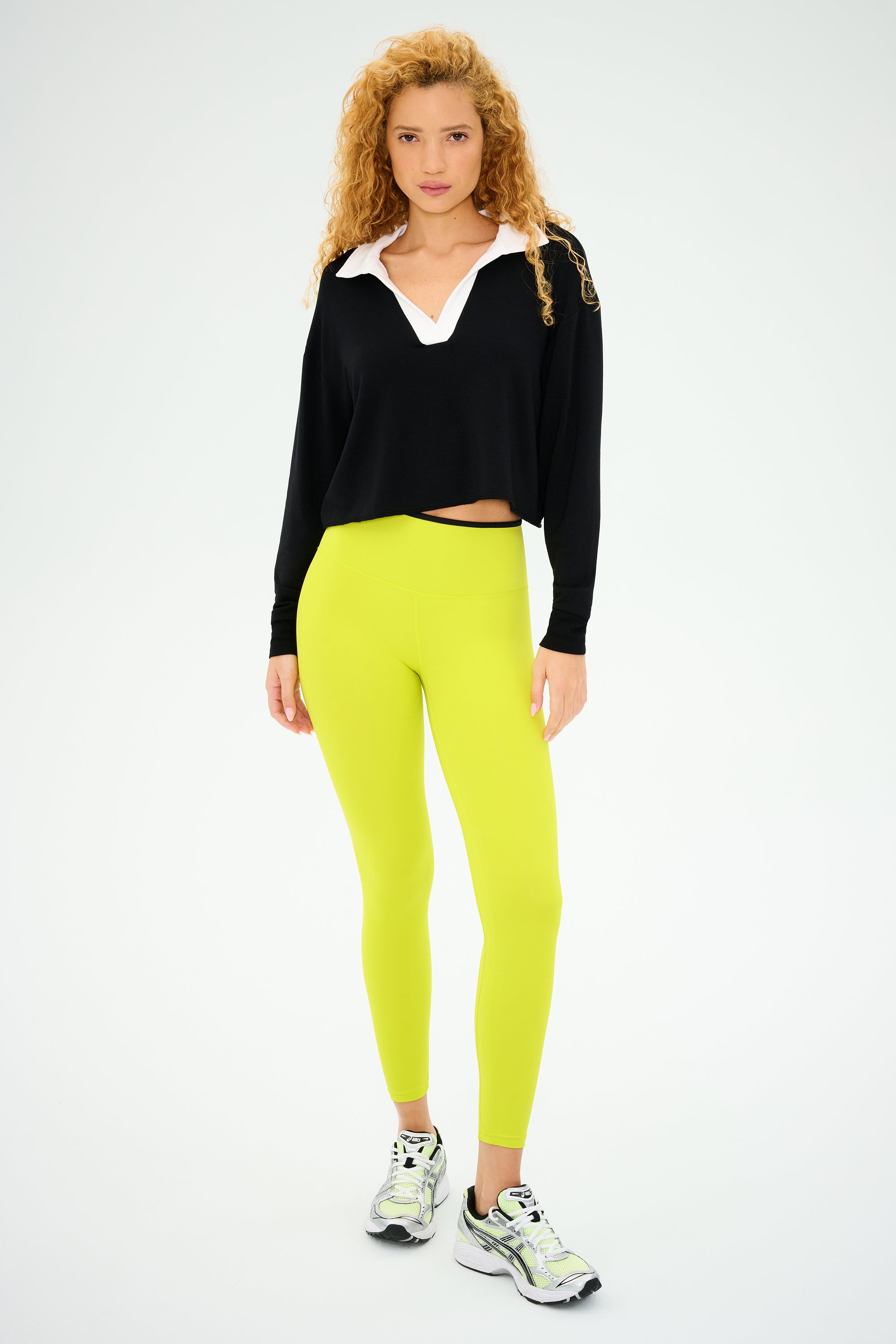 A person with curly hair stands confidently in SPLITS59's Joan Fleece Polo Sweatshirt in black/white and lime green leggings, paired with sneakers against a plain background.