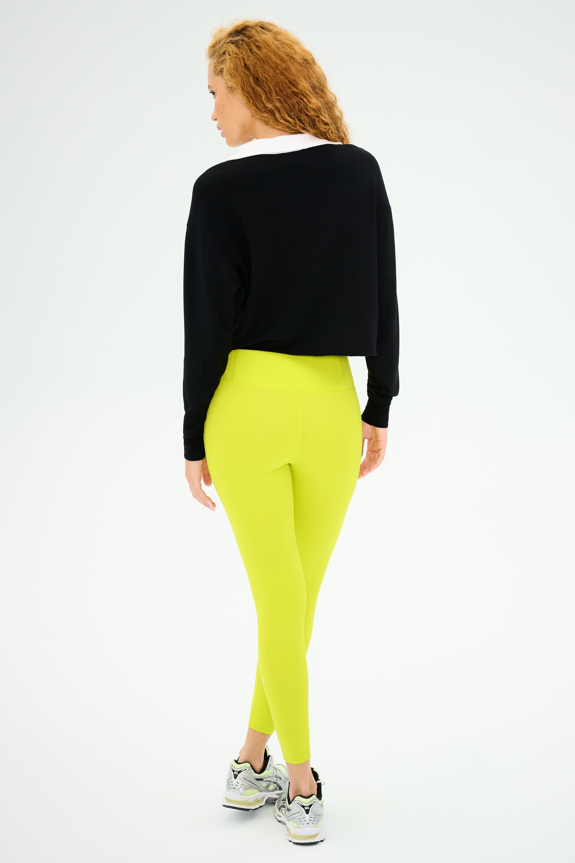 A person with curly hair wears the SPLITS59 Joan Fleece Polo Sweatshirt in black/white, neon yellow leggings made from stretch modal fabric, and sneakers while standing against a plain background with their back turned.