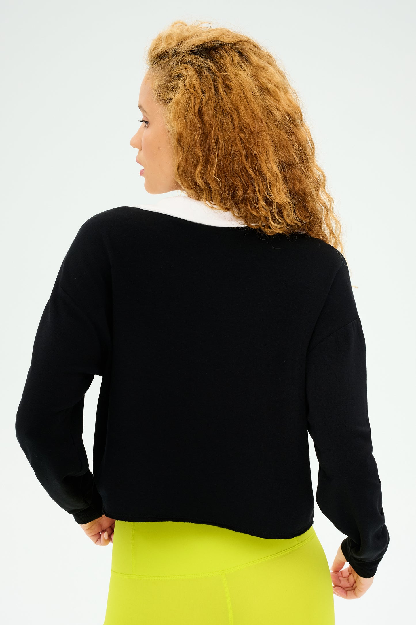 A person with curly hair is seen from behind, wearing a black Joan Fleece Polo Sweatshirt by SPLITS59 and yellow stretch modal pants.