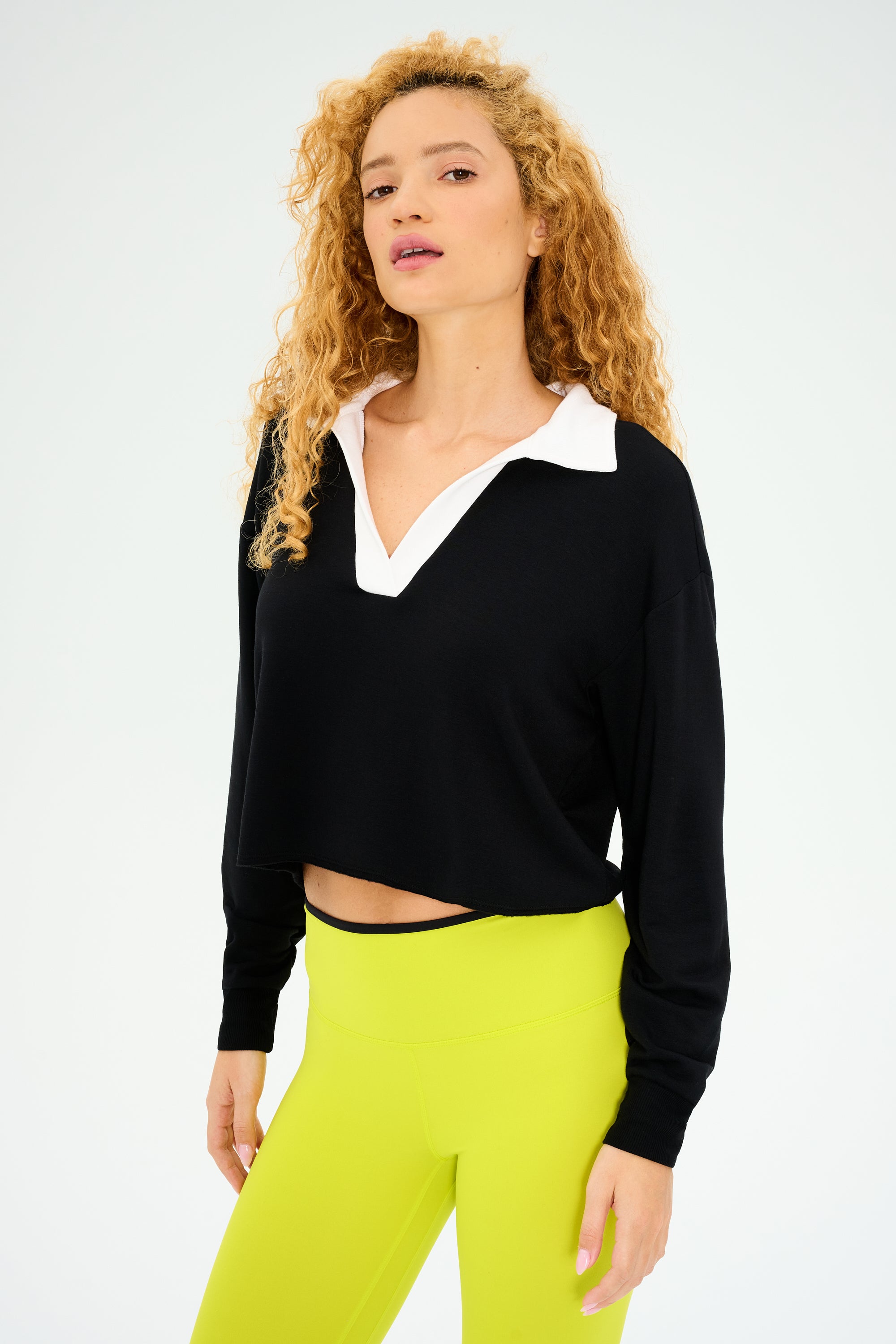 A person with curly hair wears the Joan Fleece Polo Sweatshirt in black and white by SPLITS59, paired with neon green stretch modal pants, looking at the camera.