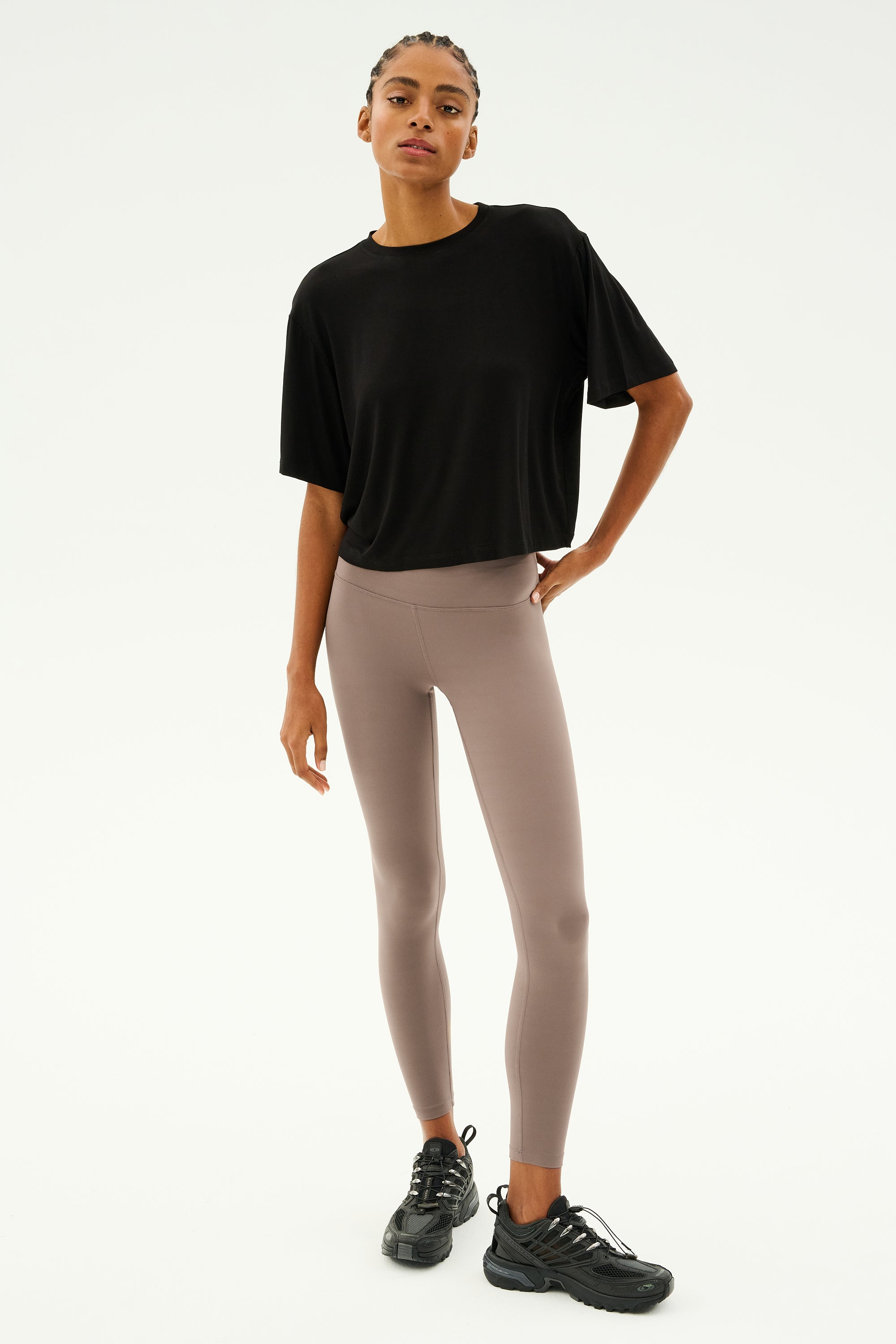 A person wearing the Djuna Oversized Jersey Tee in black from SPLITS59, paired with beige leggings and black sneakers, stands against a plain background.