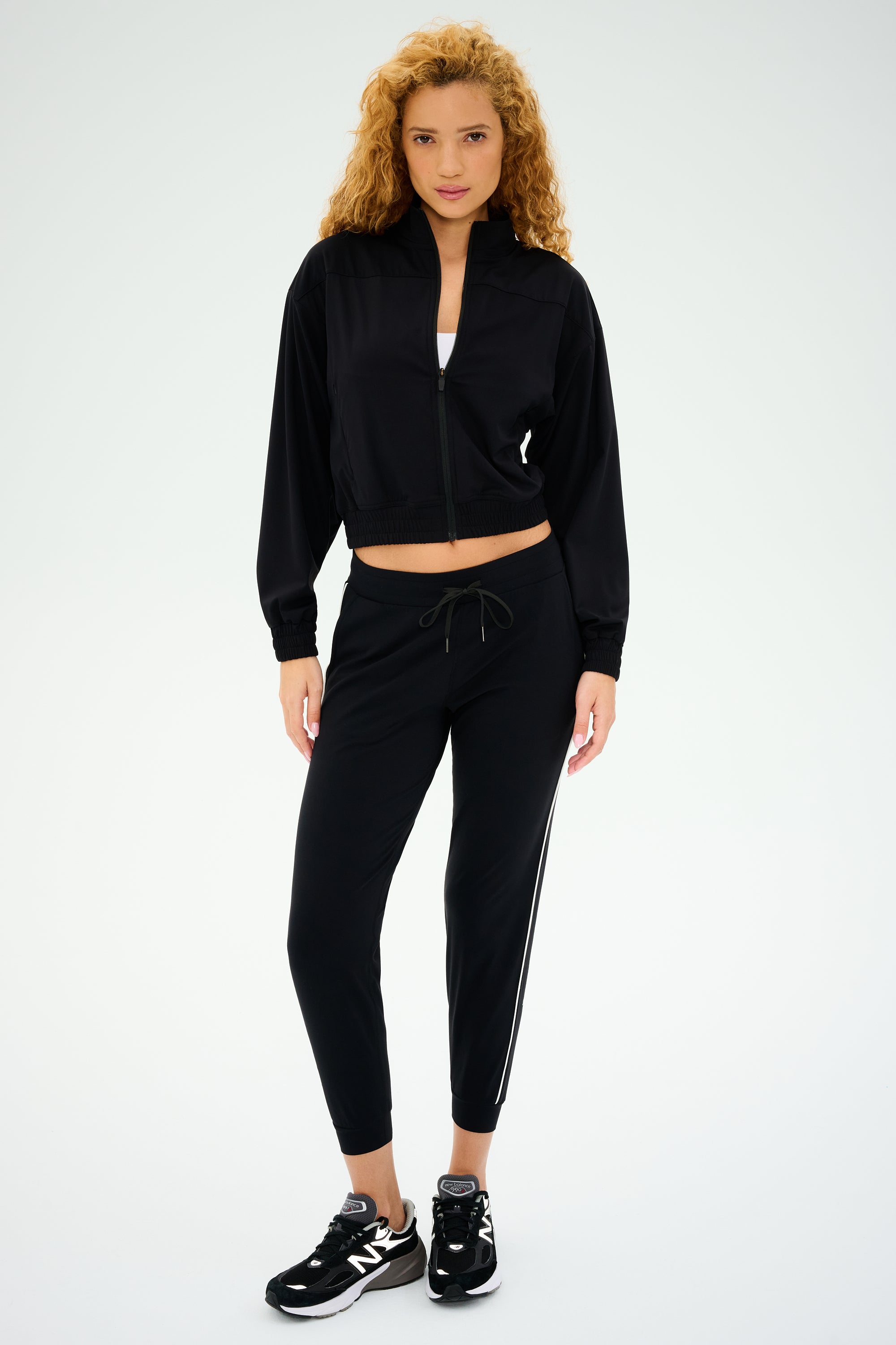 A curly-haired person models the SPLITS59 Harlowe Rigor Jacket in black, a versatile workout staple, while wearing sneakers against a plain white background.