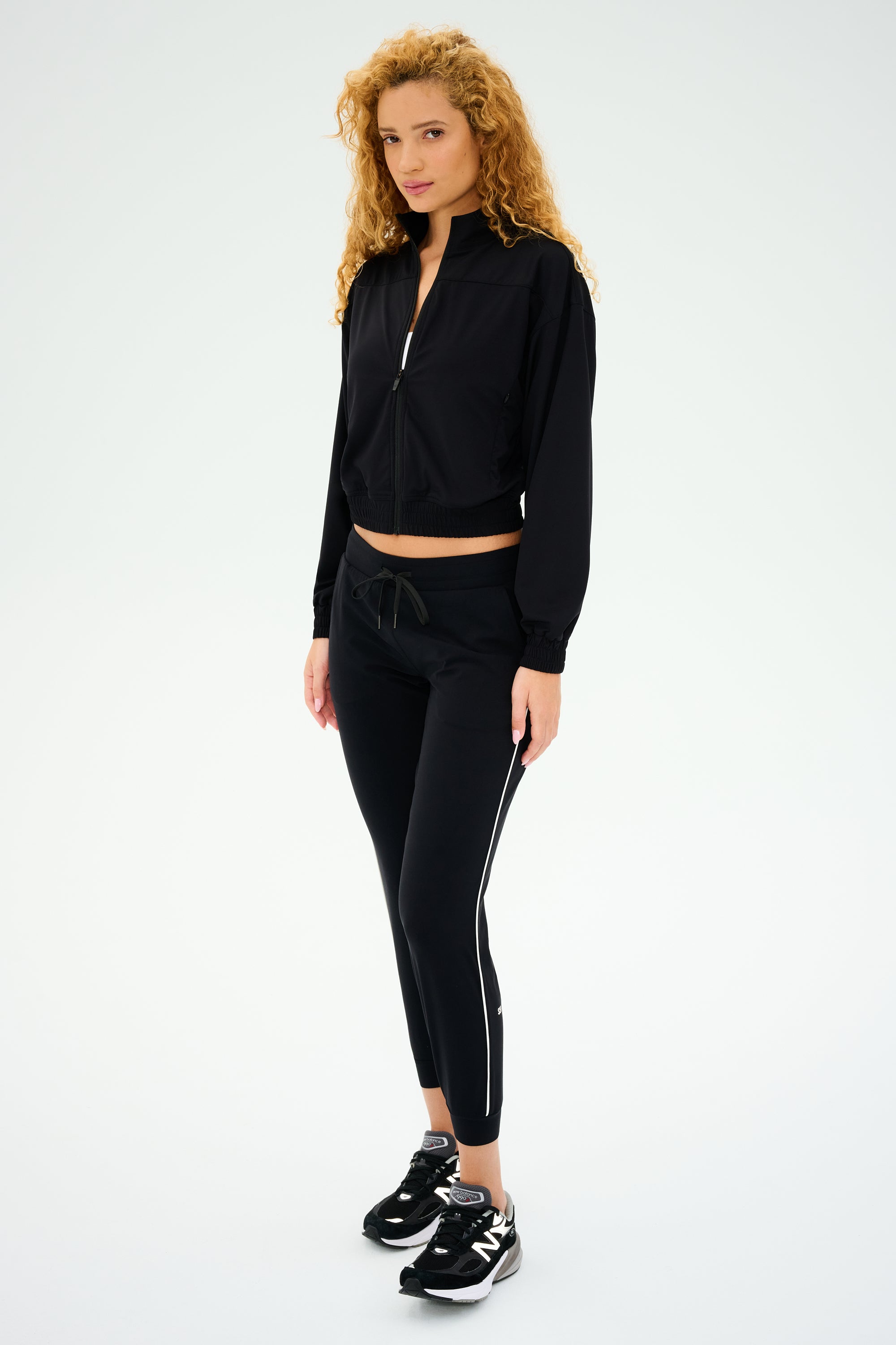 Person with curly hair in a black tracksuit and sneakers, effortlessly blending comfort and style. Their look is completed with the Harlowe Rigor Jacket - Black by SPLITS59, an all-season essential perfect for any active lifestyle against a plain backdrop.