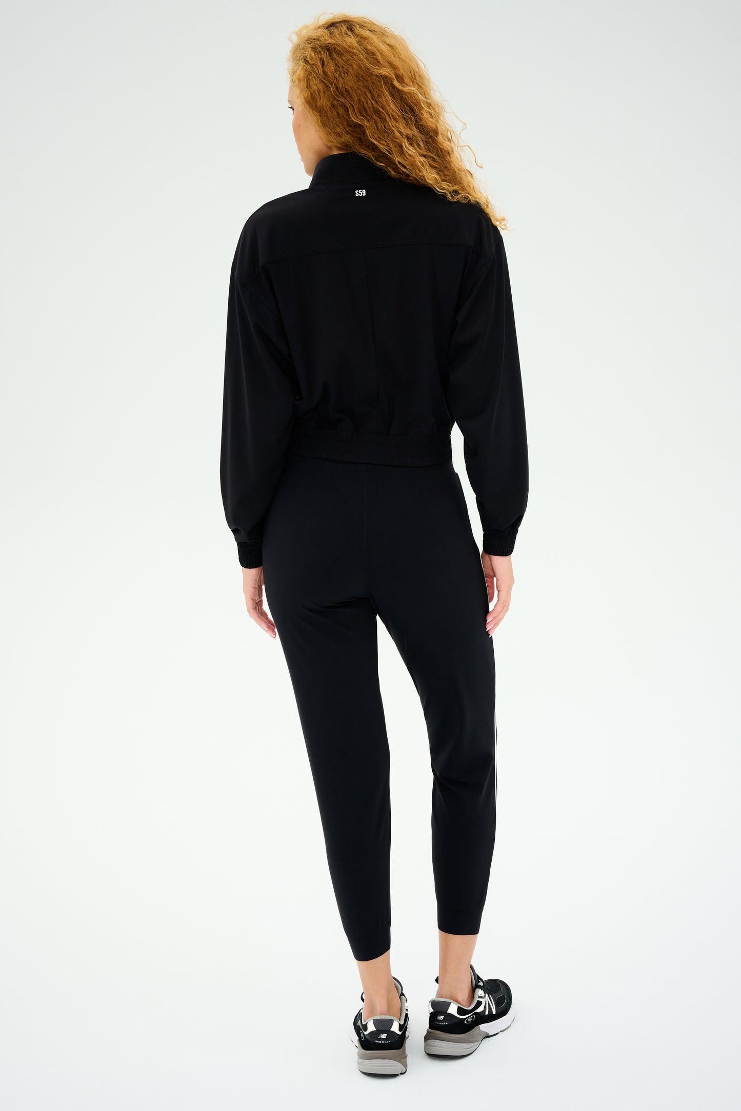 A person with curly hair, dressed in a black tracksuit and sneakers, faces away against a plain background, showcasing the SPLITS59 Harlowe Rigor Jacket in black to enhance their workout wardrobe.