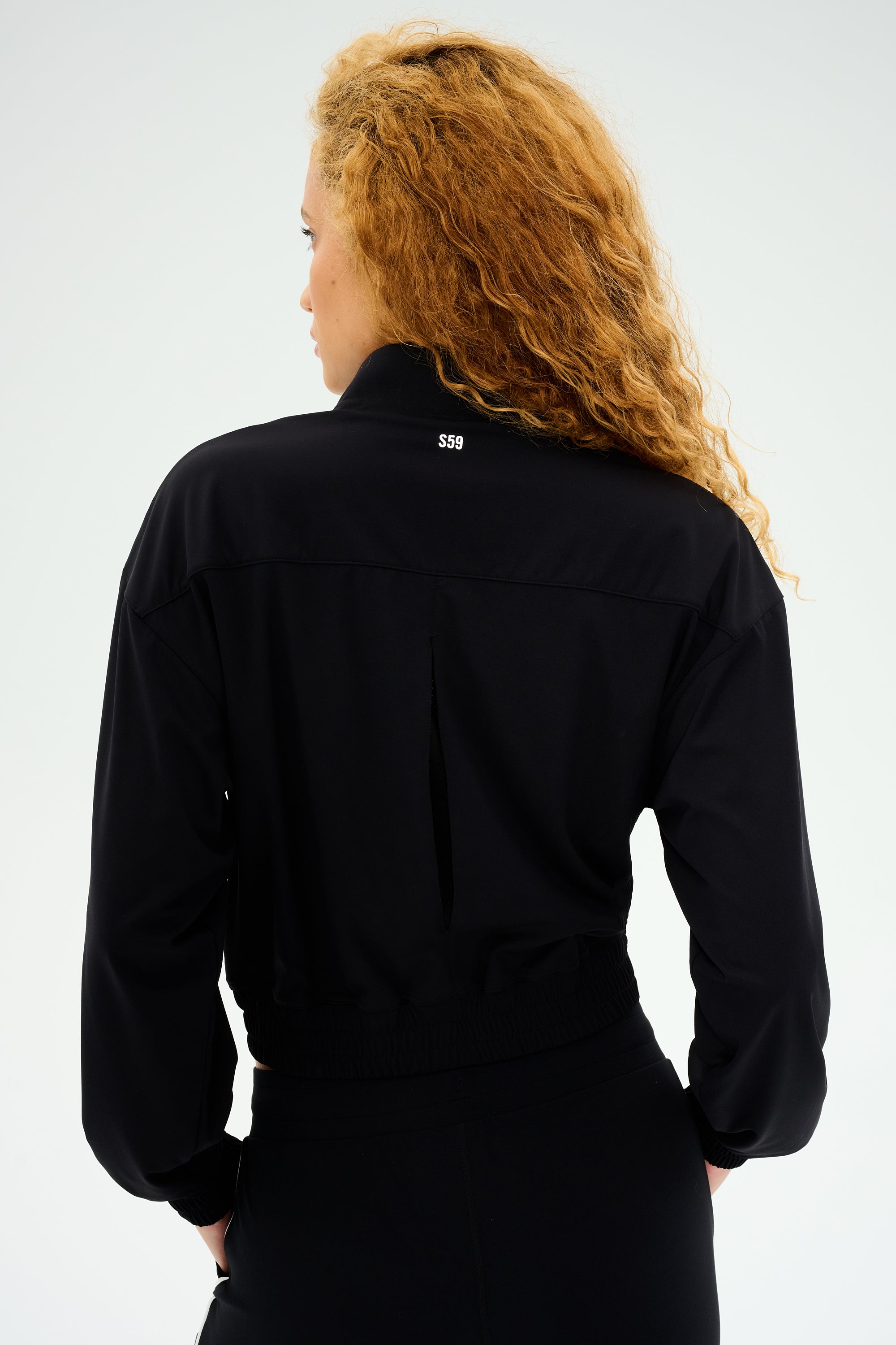 A person with long curly hair is wearing a SPLITS59 Harlowe Rigor Jacket - Black and facing away from the camera.