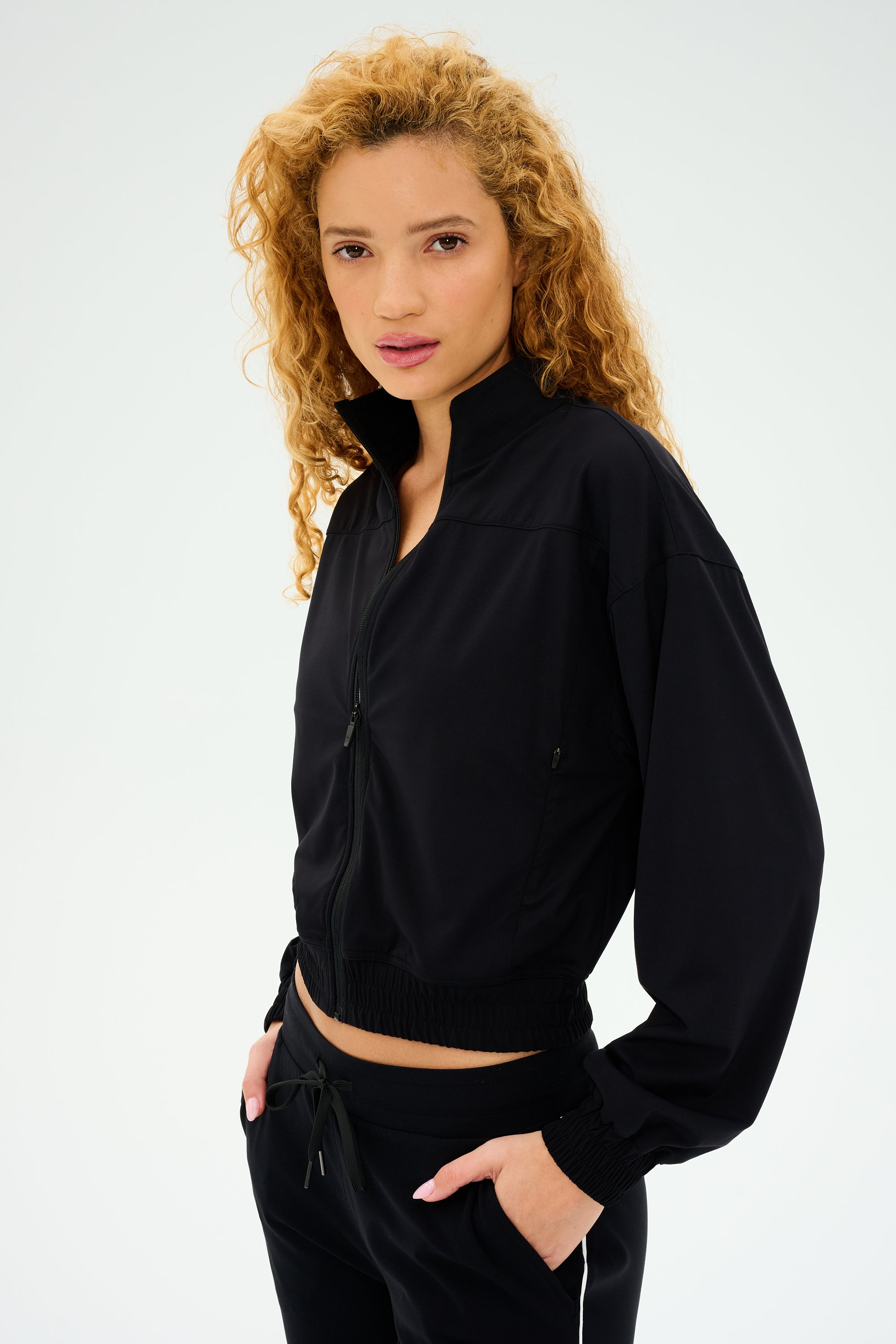 A person with curly hair stands against a plain background, wearing the SPLITS59 Harlowe Rigor Jacket - Black, paired with black pants.