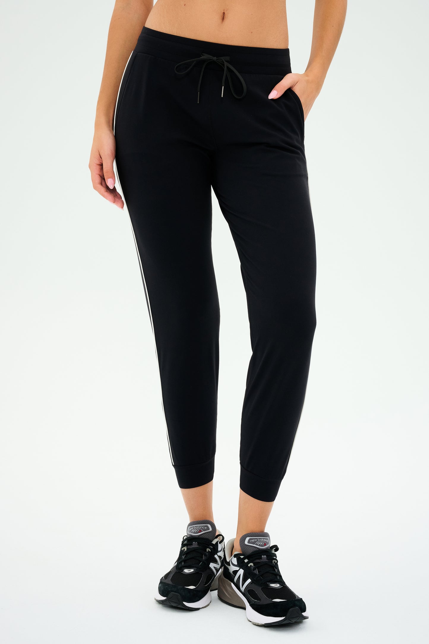 Wearing SPLITS59's Lucy Rigor Pant W/Piping in Black, a person exudes all-day comfort as they stand against a plain background, completing the look with black sneakers.