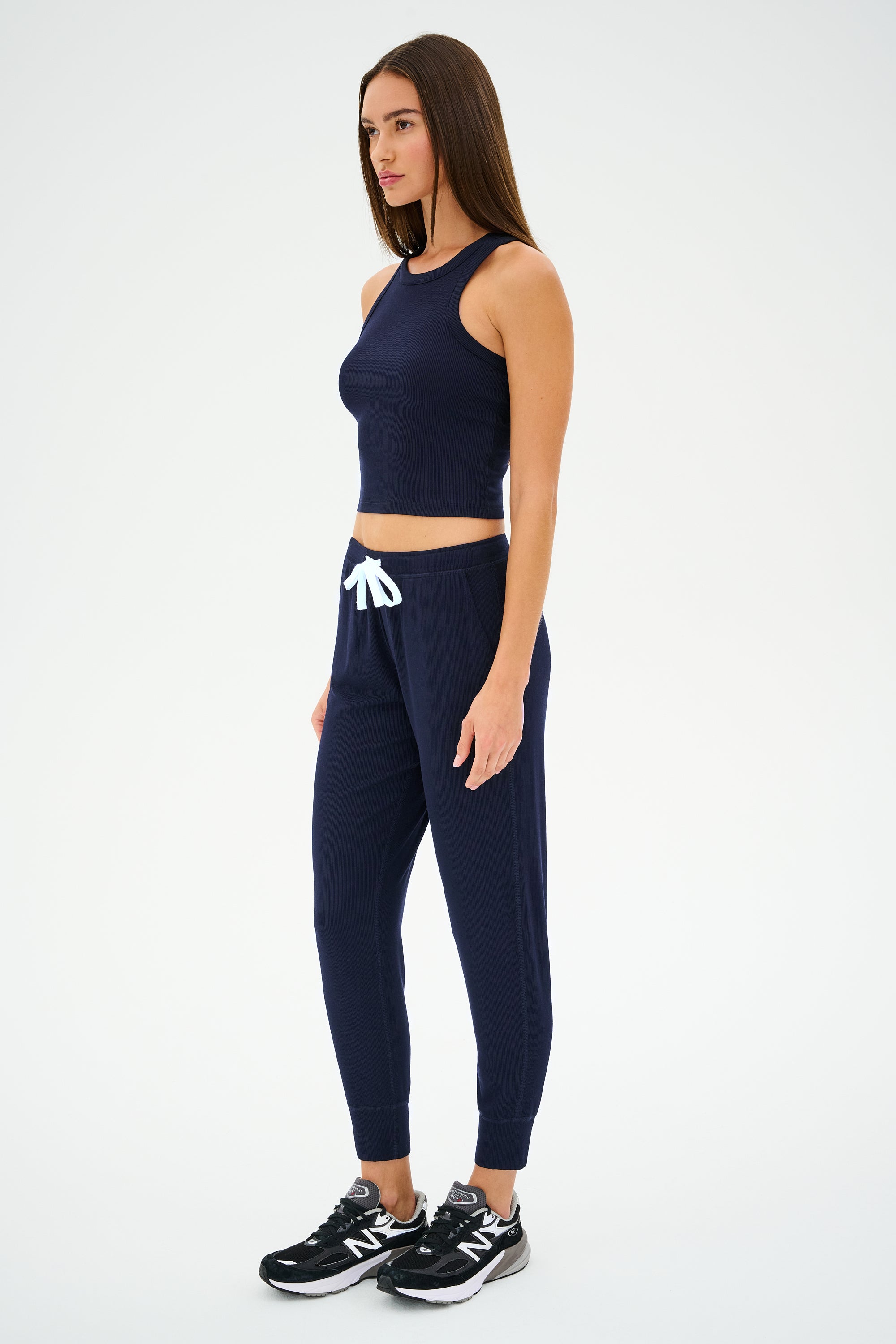 A person stands in profile wearing the Kiki Rib Crop Tank in Indigo by SPLITS59, paired with jogger pants made from baby rib fabric and black sneakers. Perfect for gym workouts, the look is set against a plain white background.