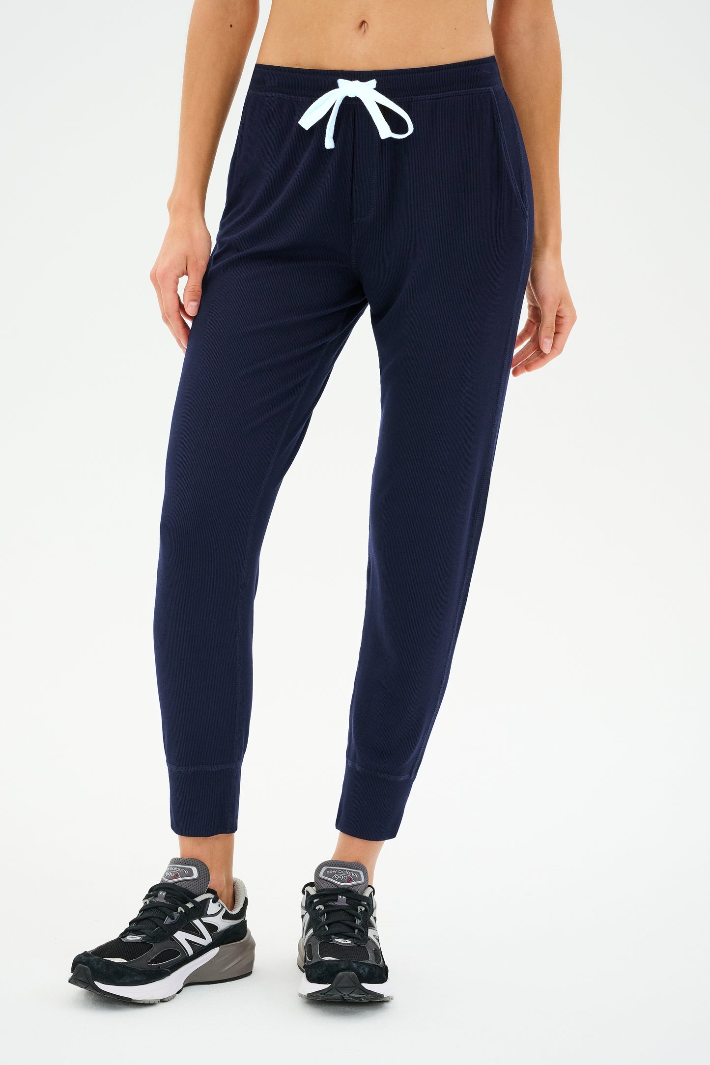 Wearing the Kiki Rib 7/8 Sweatpant in Indigo from SPLITS59, a person epitomizes effortless comfort with its navy ribbed texture and white drawstring. They complete the look with black and white sneakers.