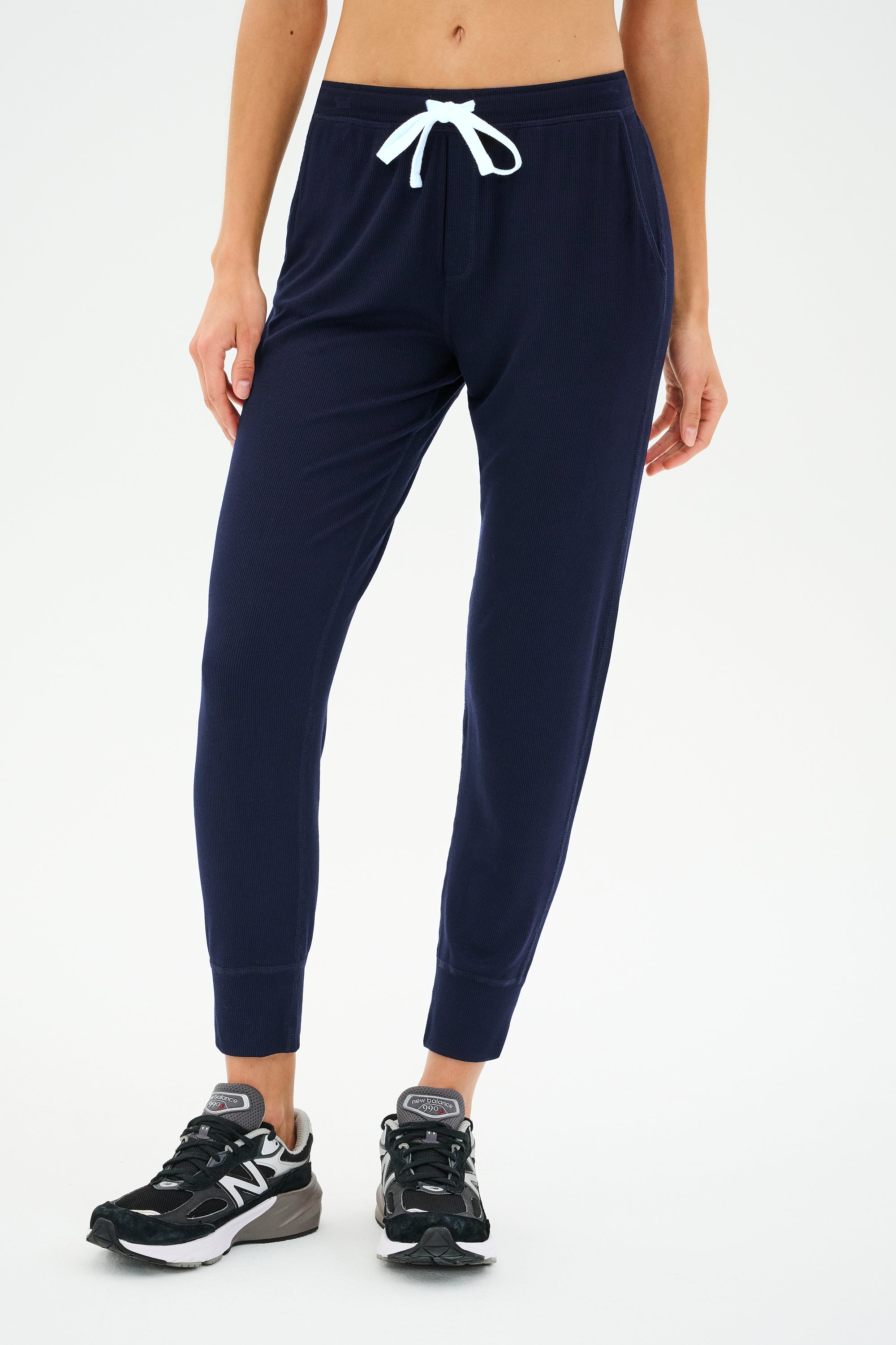 Wearing the Kiki Rib 7/8 Sweatpant in Indigo from SPLITS59, a person epitomizes effortless comfort with its navy ribbed texture and white drawstring. They complete the look with black and white sneakers.