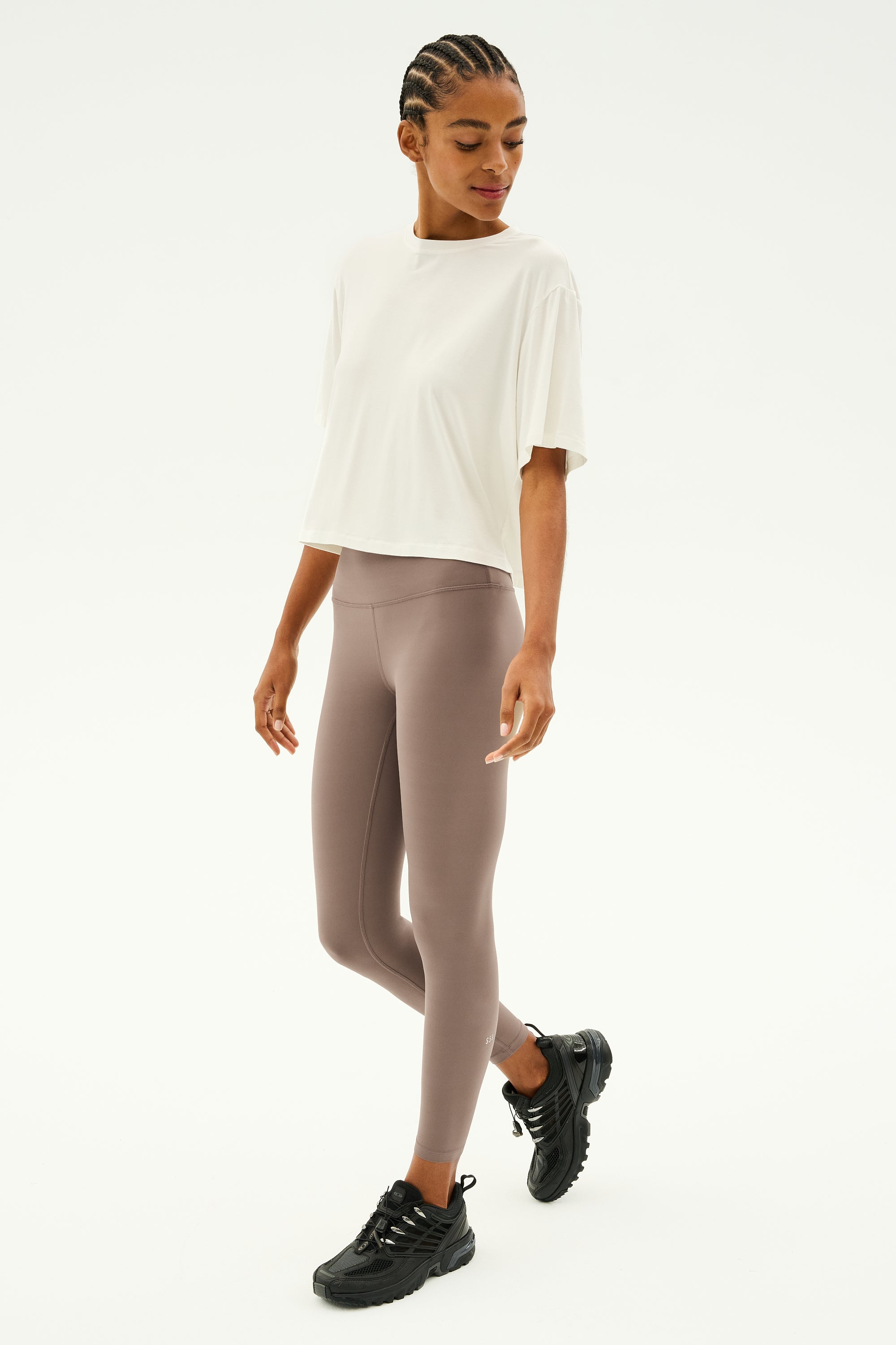 A person in a white Djuna Oversized Jersey Tee by SPLITS59, paired with taupe leggings and black sneakers, stands ready for outdoor workouts against a plain background.