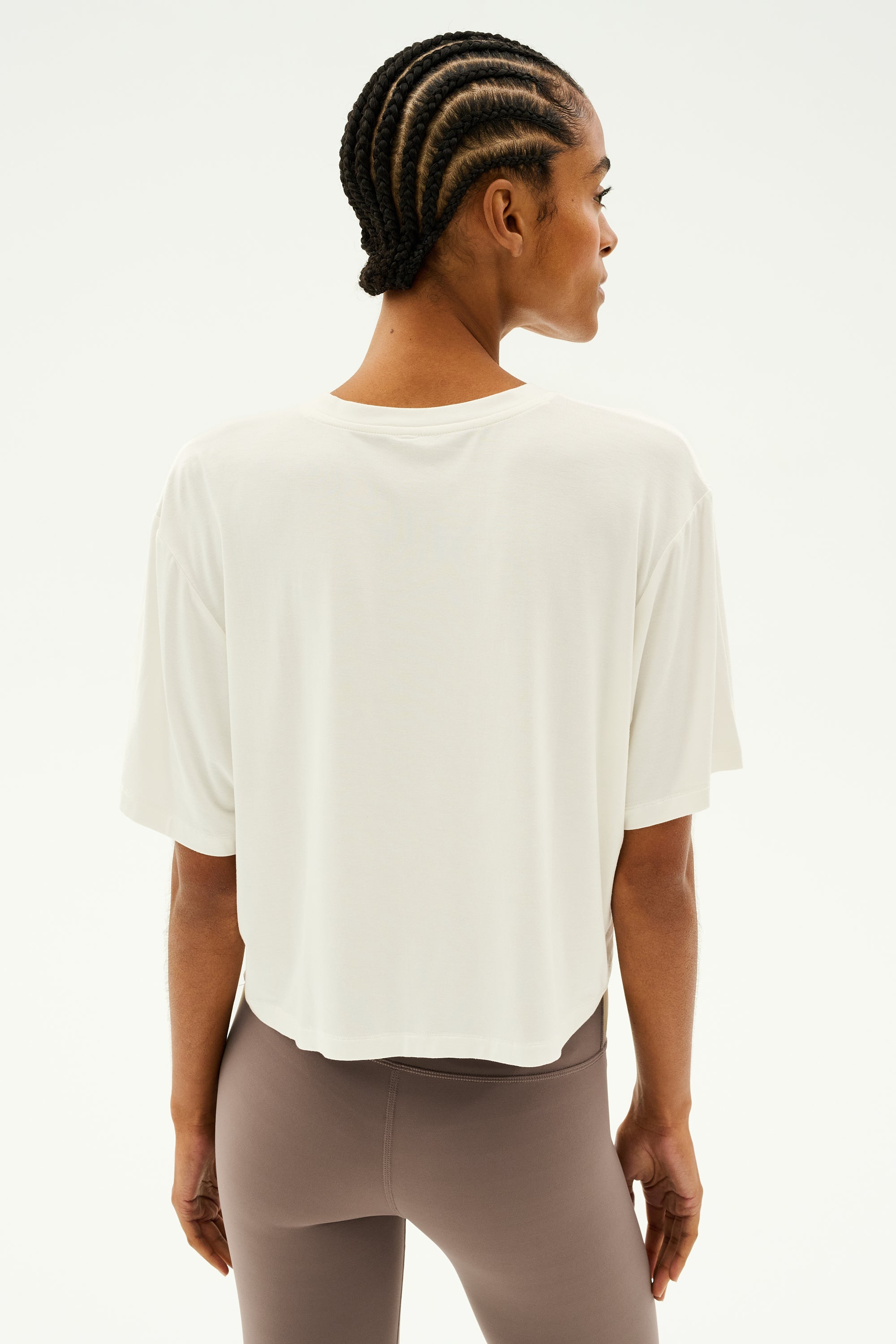 A person with braided hair wears the Djuna Oversized Jersey Tee in white by SPLITS59 and beige leggings, standing with their back to the camera against a plain background, perhaps ready for outdoor workouts.