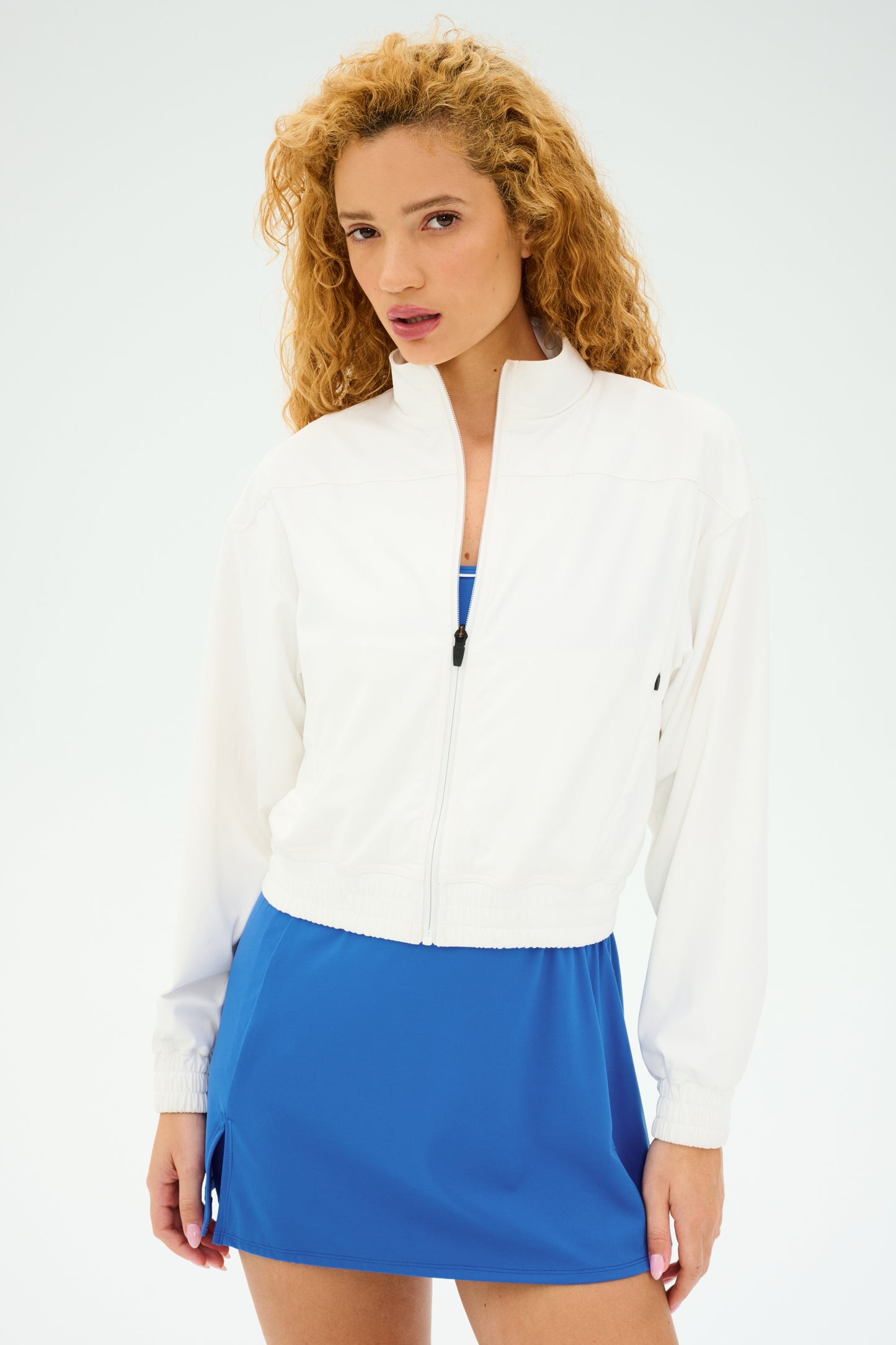 With curly hair, a person stands against a plain backdrop wearing the Harlowe Rigor Jacket in white from SPLITS59, which adds a bomber style flair to their blue dress.