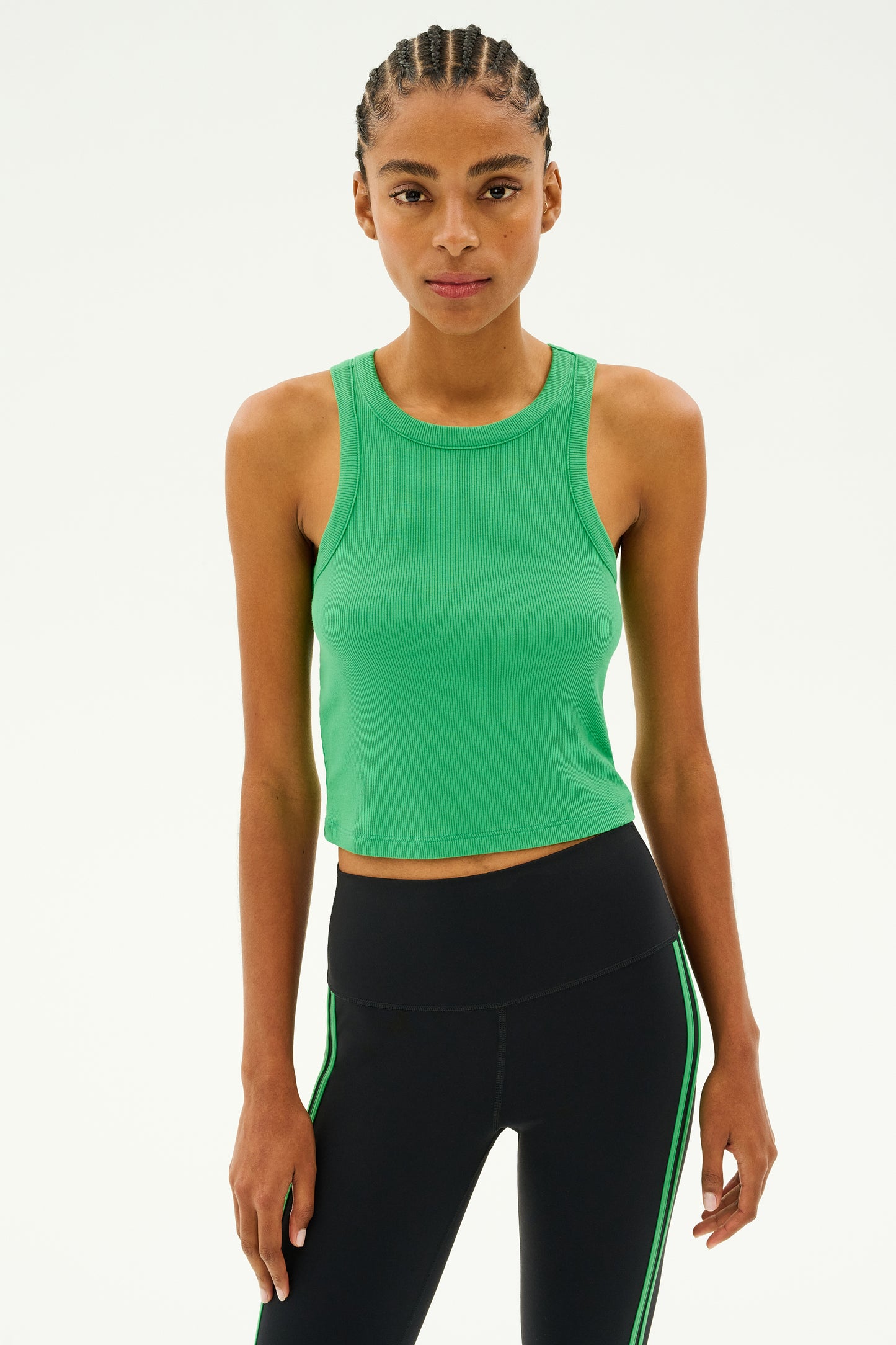 A person dressed in the Kiki Rib Crop in Grass by SPLITS59 and paired with black leggings featuring green stripes is standing against a plain background.