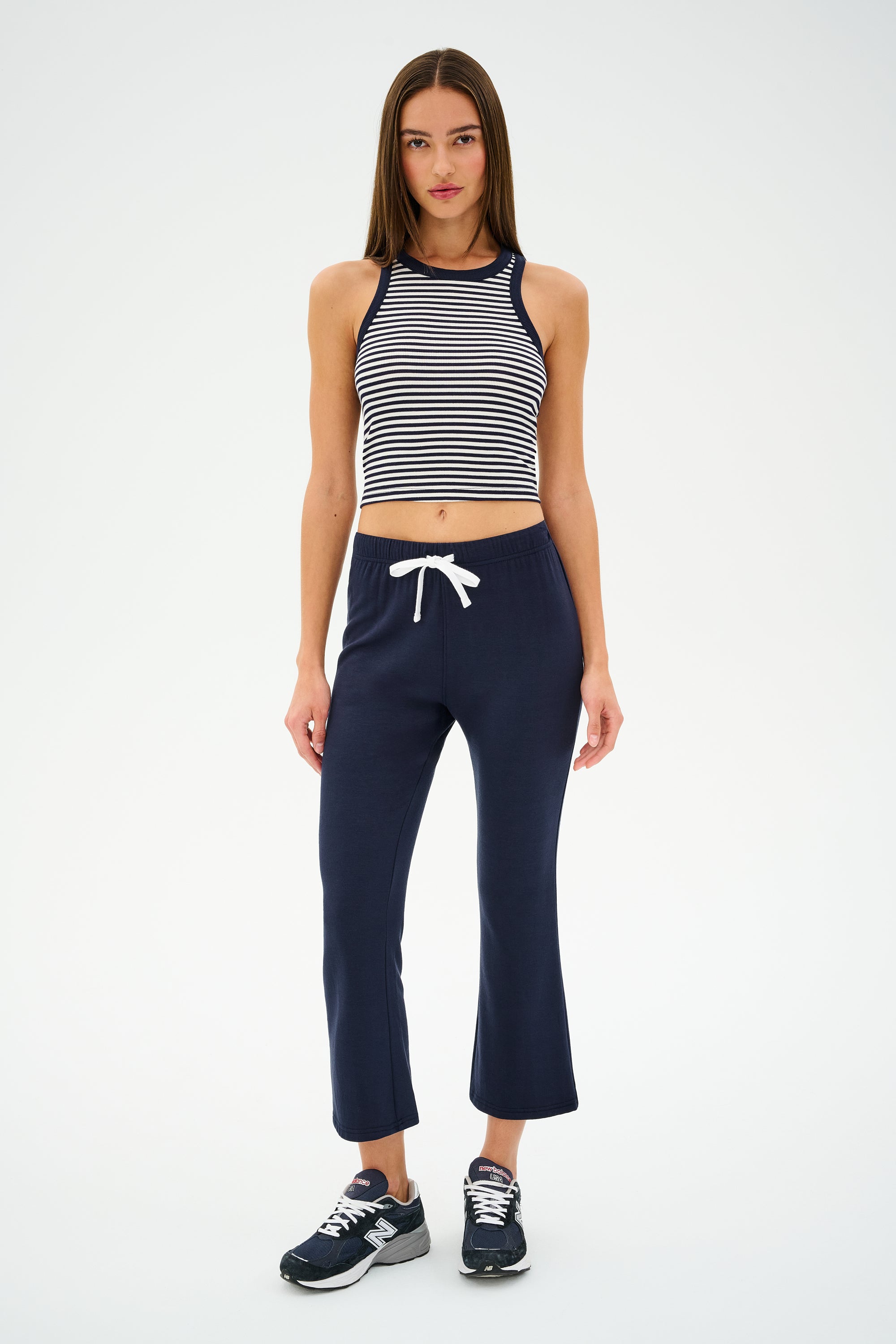 Wearing a SPLITS59 Kiki Rib Crop Tank in white and indigo stripes with navy sweatpants and black sneakers, a woman stands confidently against a plain white background, ready for her gym workouts.