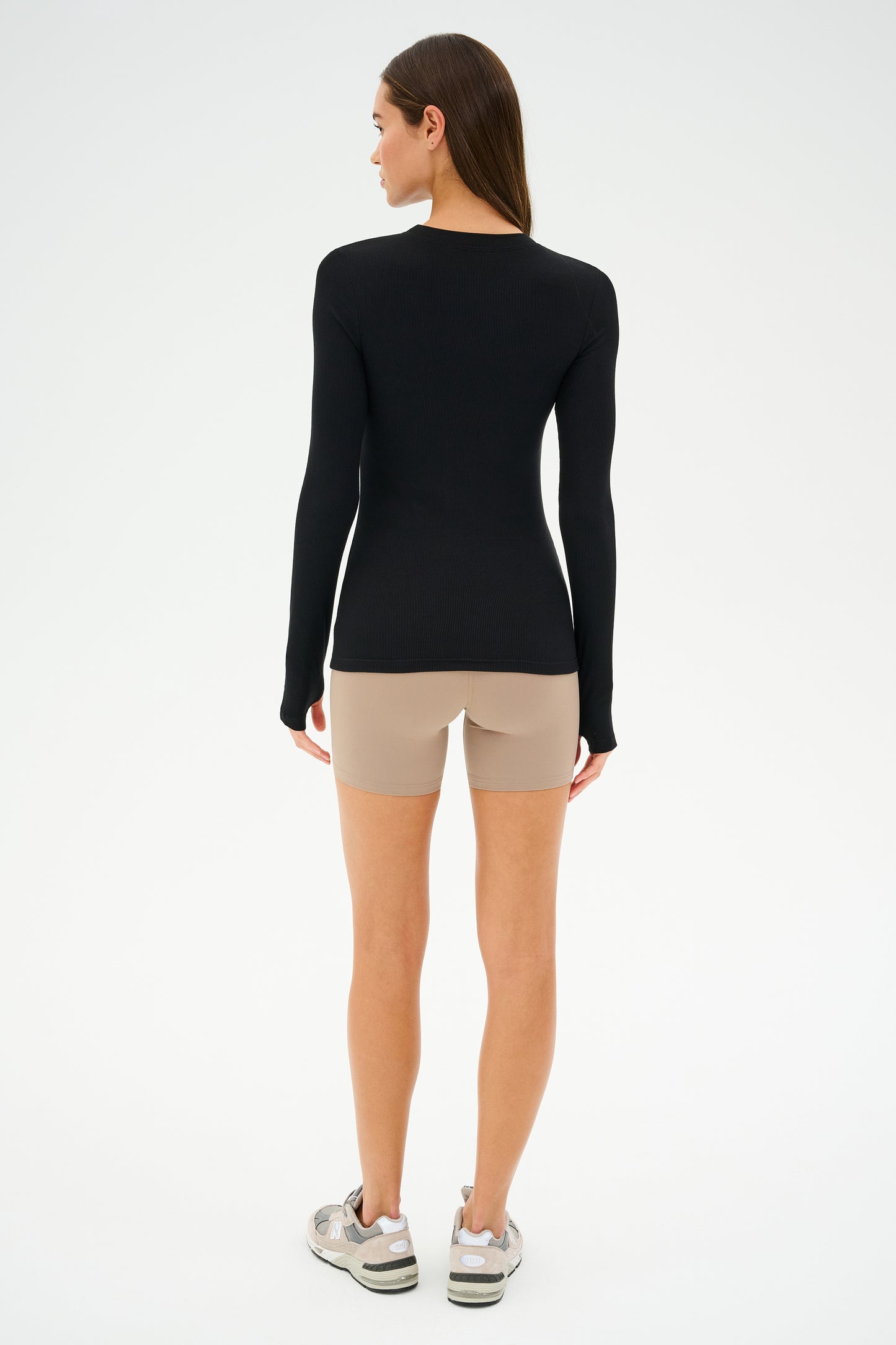 Facing away from the camera, a person wears SPLITS59's Airweight High Waist Short in Latte with a black long-sleeve top and sneakers, reflecting the simplicity and focus of a Pilates session against a plain background.