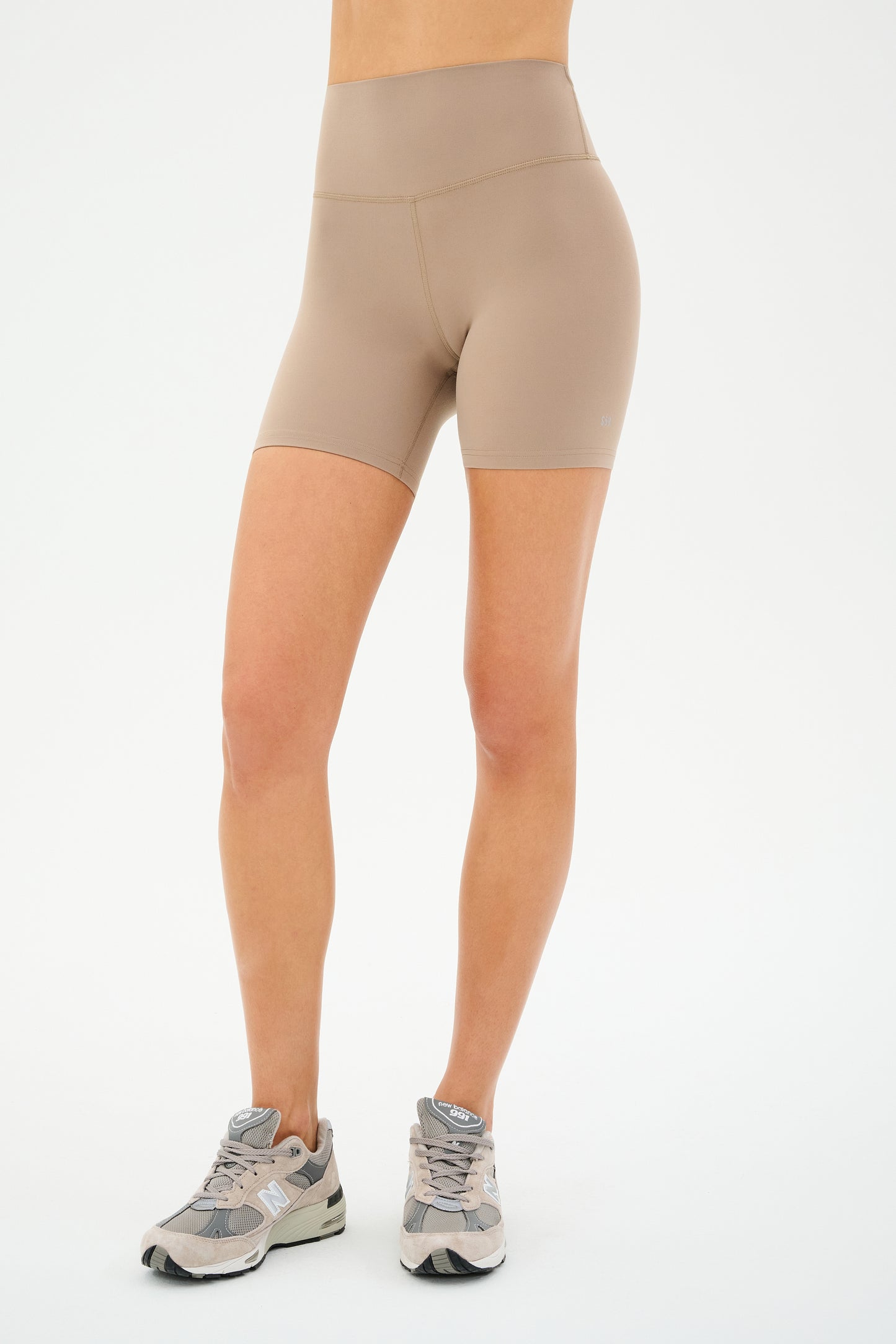 A person stands against a plain white background, wearing SPLITS59's Airweight High Waist Short in Latte and gray athletic shoes.