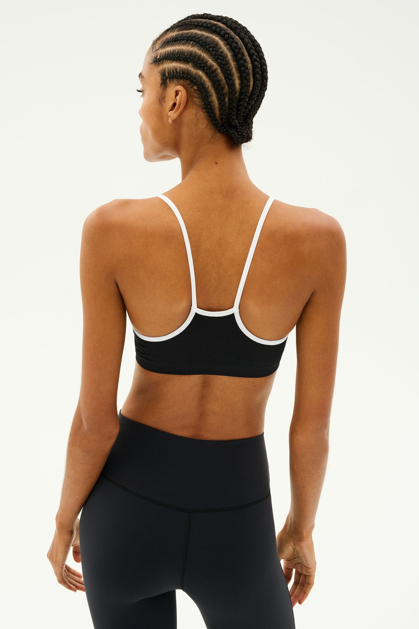 A person wearing the Loren Seamless Bra in Black/White by SPLITS59 is shown from the back, styled in black athletic wear. Their braided hair accentuates the outfit, which is crafted with chafe-free fabric for optimal gym workouts. They stand confidently against a plain background.