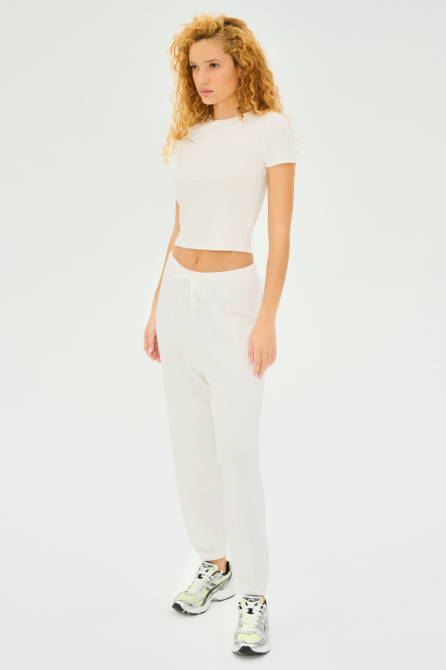 A person with curly hair stands against a plain backdrop, wearing a casual style featuring the Jennie Jersey Tee in white by SPLITS59, paired with matching white pants and sleek sneakers.