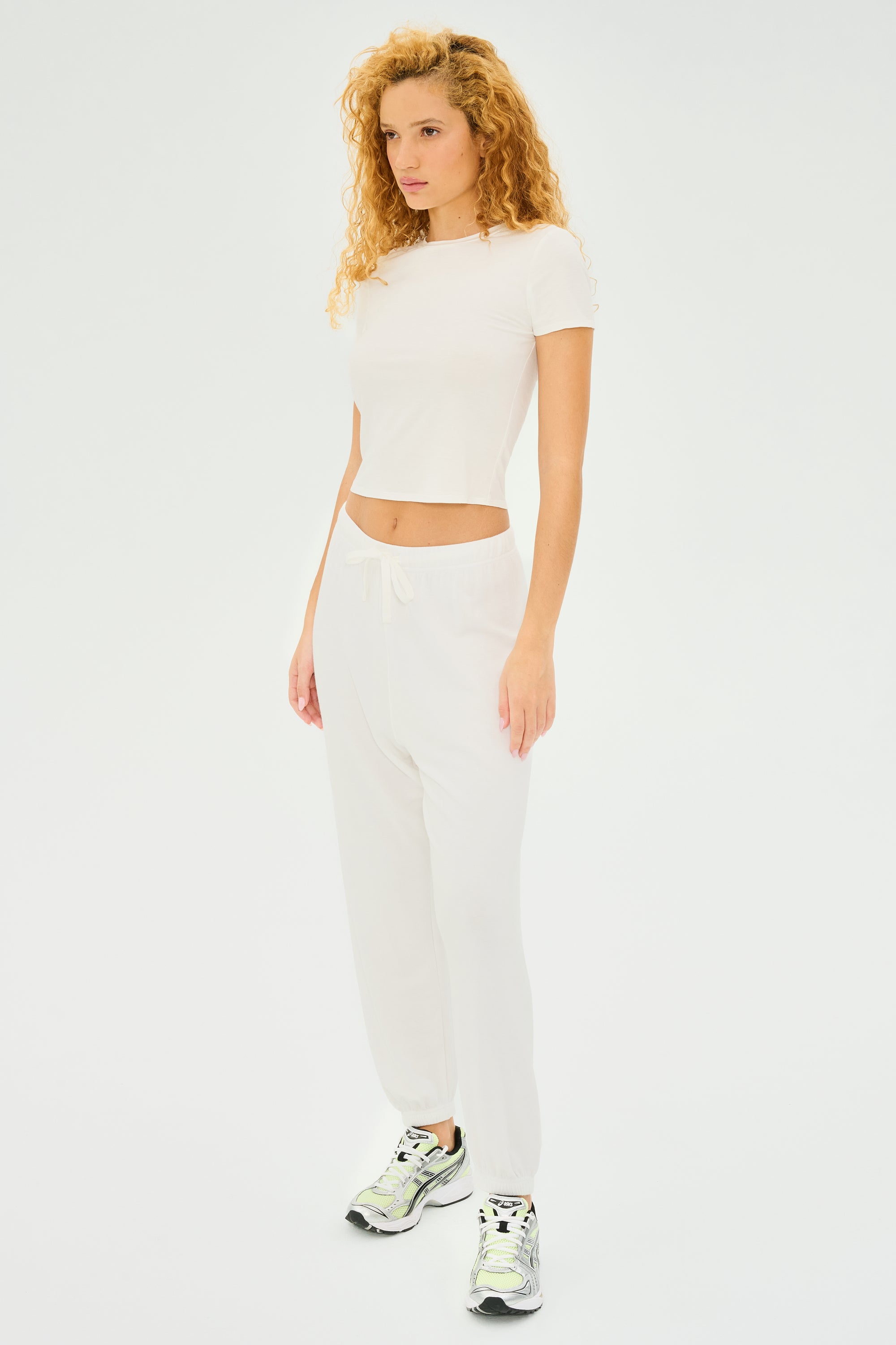 A person with curly hair stands against a plain backdrop, wearing a casual style featuring the Jennie Jersey Tee in white by SPLITS59, paired with matching white pants and sleek sneakers.