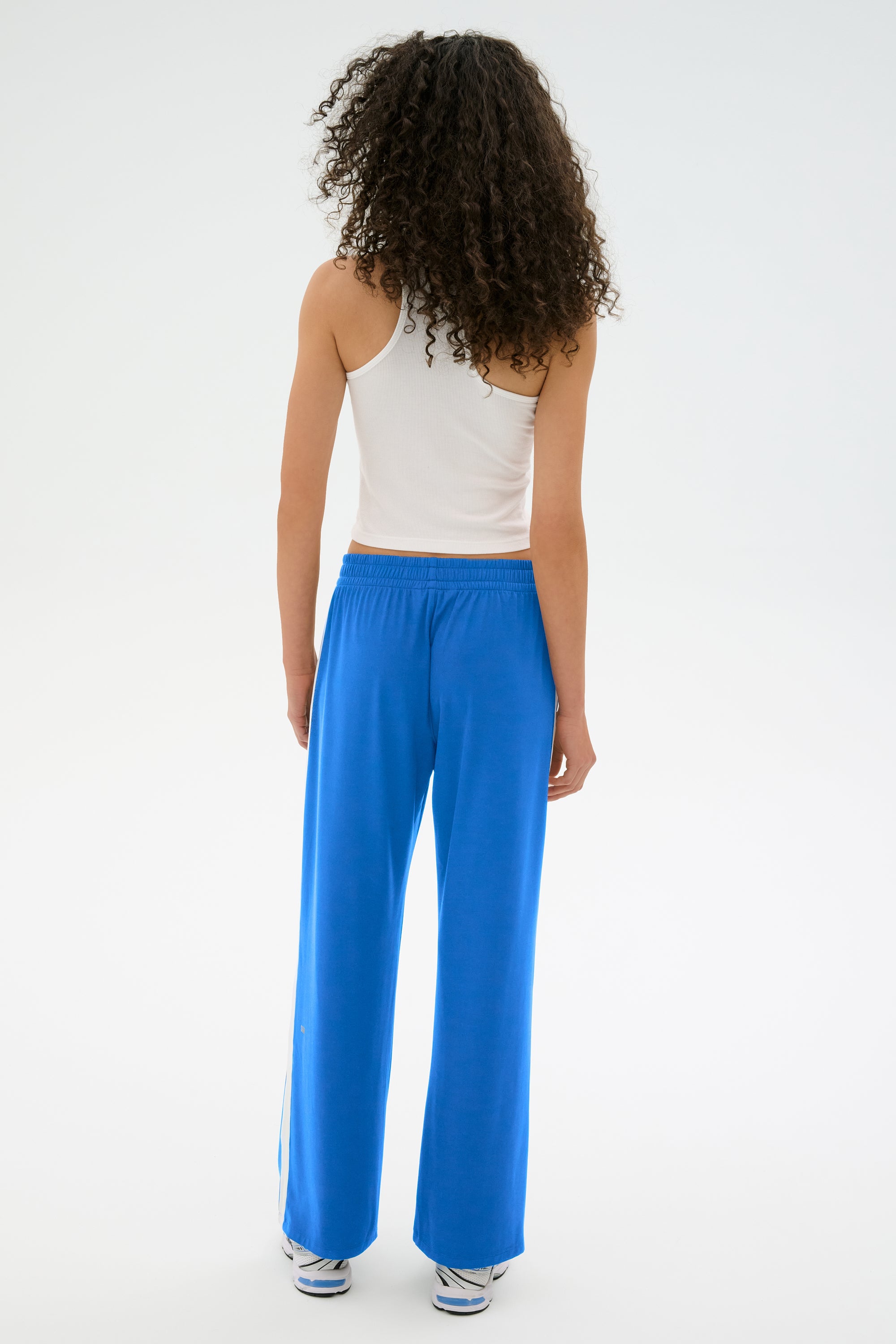 A person with curly hair is seen from behind, wearing a white tank top and SPLITS59's Maxie Rigor Track Pant in Classic Blue/White, achieving a stylish and relaxed look.