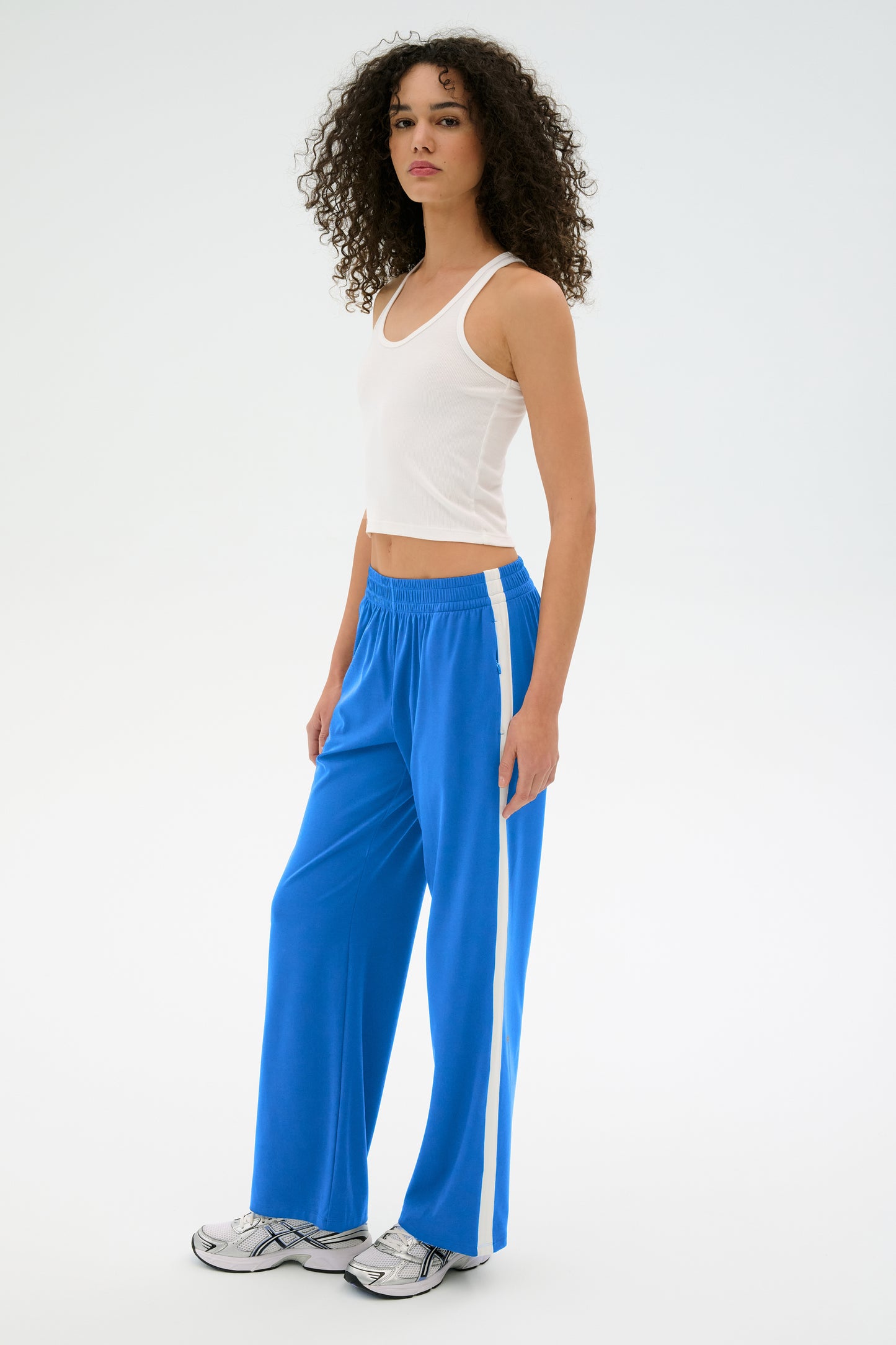 A curly-haired person confidently stands against a white backdrop, wearing a white tank top and SPLITS59's Maxie Rigor Track Pant in Classic Blue/White.