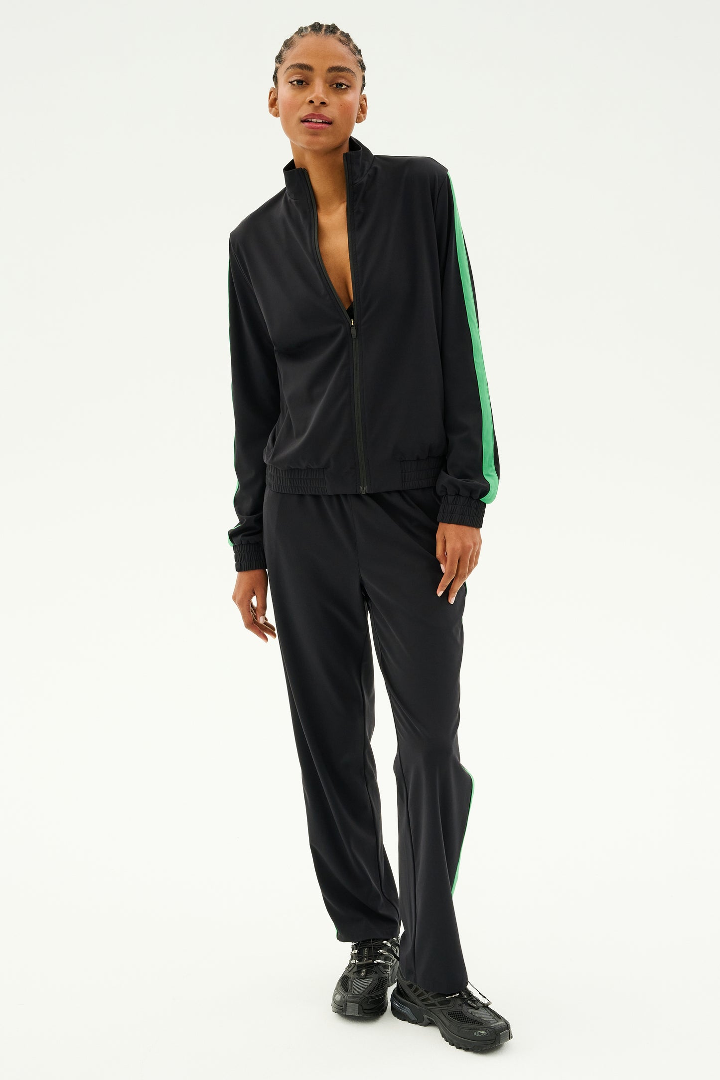 A person wearing the SPLITS59 Max Rigor Track Pant in Black/Grass, featuring a sporty fit with green stripes and wide-leg pants, stands against a plain background, complemented by sleek black shoes.